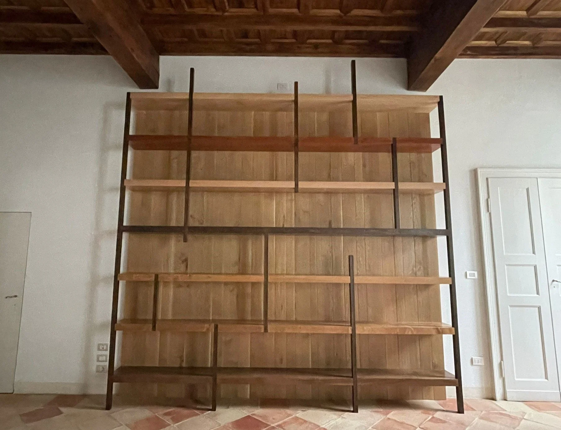 Scaffale vuoto di legno e metallo in una stanza con pareti bianche e pavimento di piastrelle in tonalità terracotta.