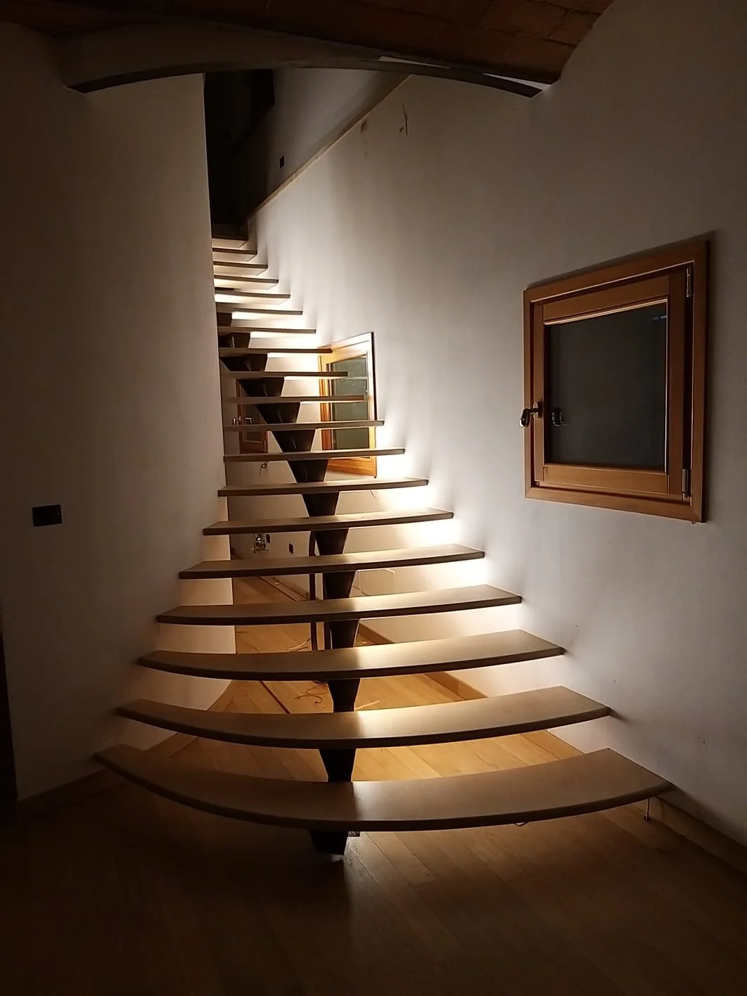 Scala a chiocciola in legno con illuminazione soffusa, vista dall'interno di una casa moderna.