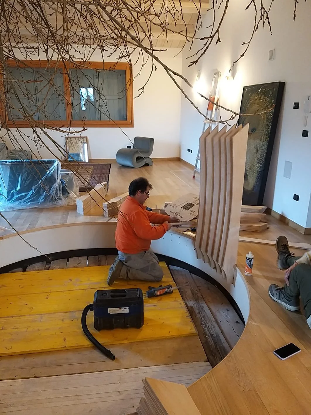 Uomo che lavora a un progetto di falegnameria in un interno moderno, con pannelli di legno e strumenti da lavoro, in un ambiente illuminato con mobili e grandi finestre.