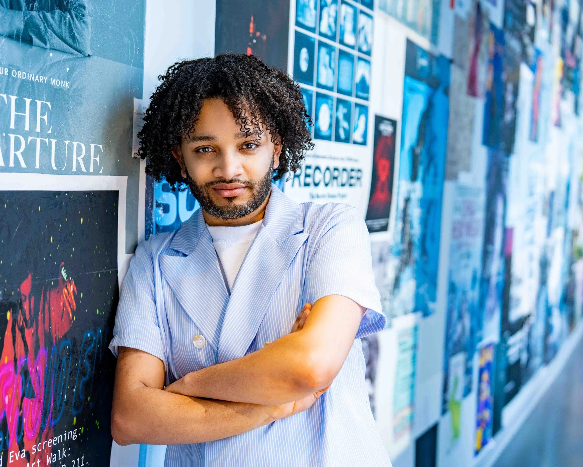 A man with curly hair and a beard leaning against a wall with posters, crossing his arms and looking at the camera.