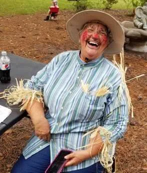 Woman dressed in a striped shirt and a large sun hat, with face paint and straw accessories, smiling and sitting outdoors.