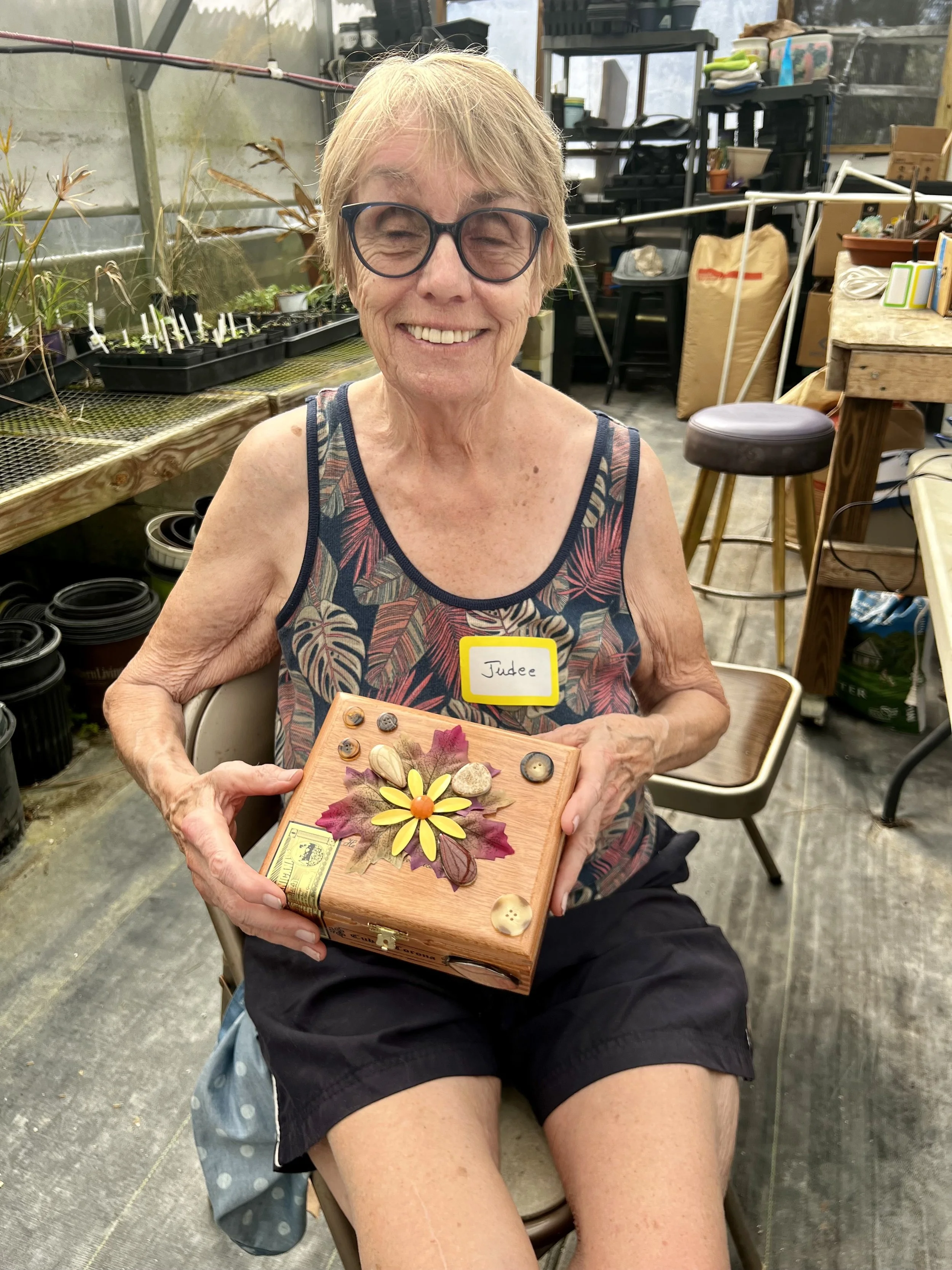 An elderly woman with short blonde hair, glasses, and a sleeveless top is sitting in a greenhouse or gardening workshop, smiling and holding a decorated wooden box with various natural elements. She wears a yellow name tag labeled 'Judee'.