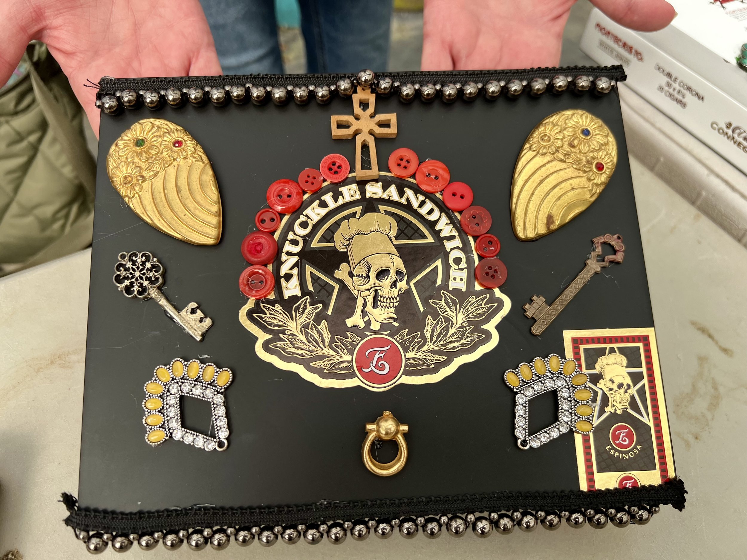 Black box decorated with badge featuring a skull, red buttons, keys, gold and silver embellishments, and a cross in the center.