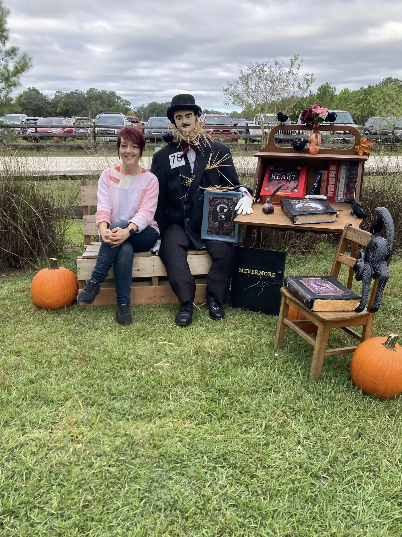 A woman sitting on a bench next to a mannequin dressed as Edgar Allan Poe, both are outdoors with pumpkins and autumn decorations around them.