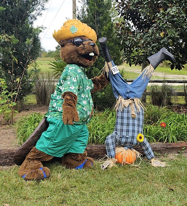 Costumed bear character dressed in summer clothes standing outdoors with a garden and trees in the background, holding a sign numbered 93. The bear poses next to a scarecrow dressed in plaid clothing with a sunflower on its waist and a pumpkin at its