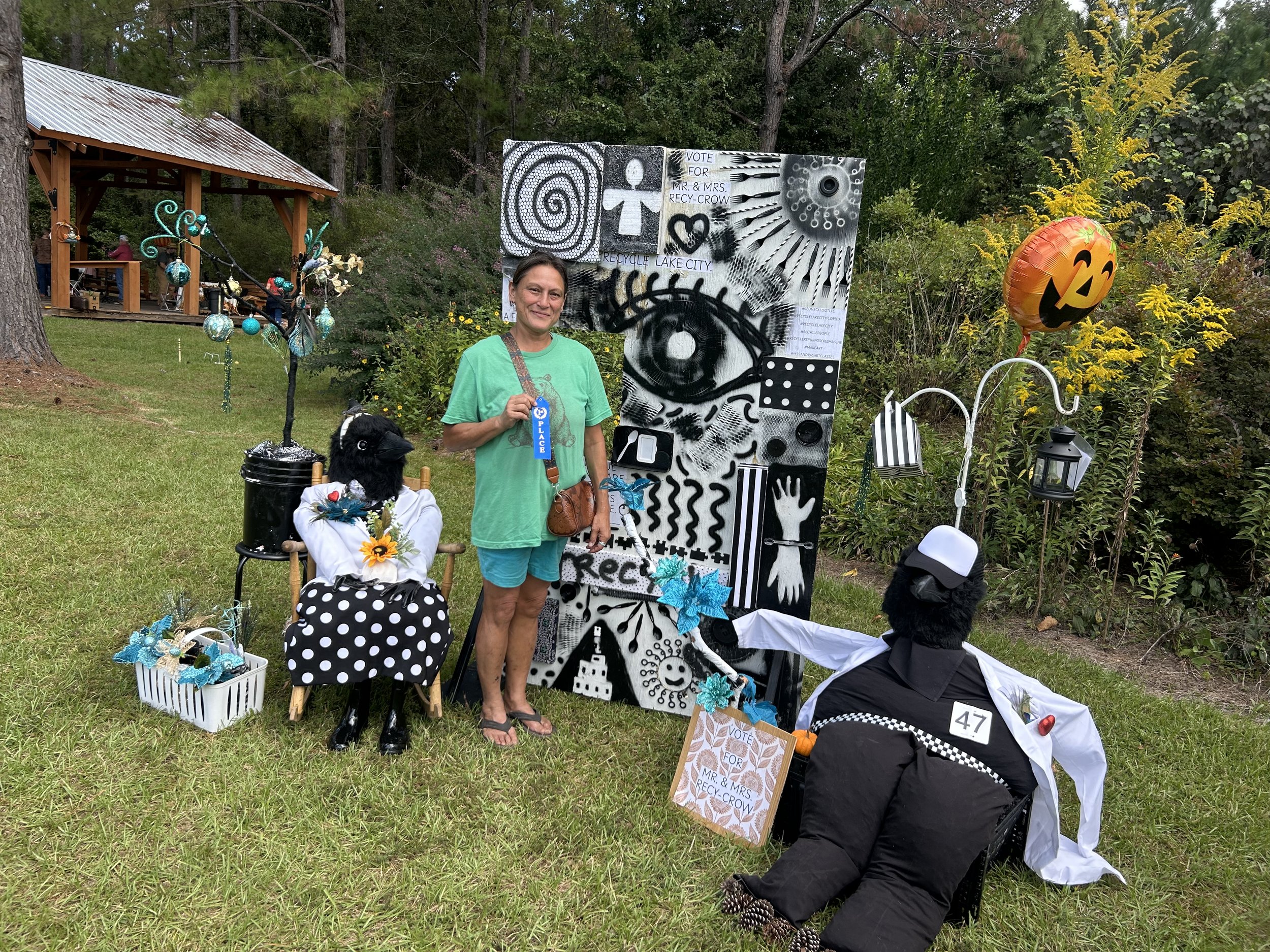 A woman stands outdoors in front of Halloween-themed decorations, including large black and white figures with police theme, a pumpkin balloon, and a decorative tree with blue ornaments. The scene includes a picnic table in the background, trees, and