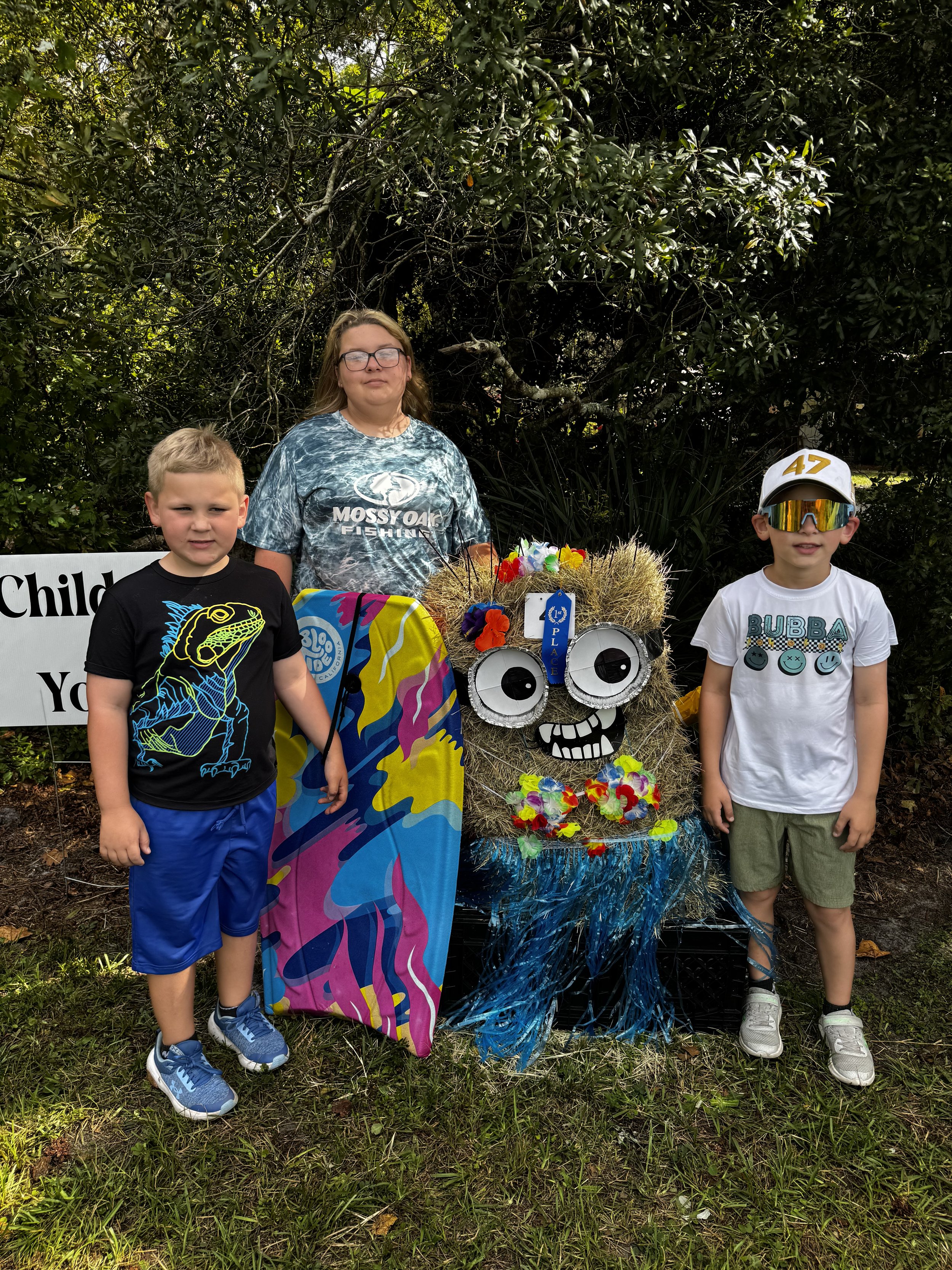 Three children standing next to a colorful decorative display with a face made of various craft materials, flowers, and tinsel, outdoors in front of leafy bushes.