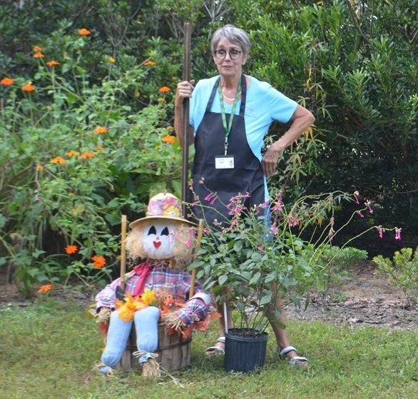 A woman standing outdoors surrounded by green plants and flowers, holding a gardening tool with a scarecrow decoration sitting beside her.