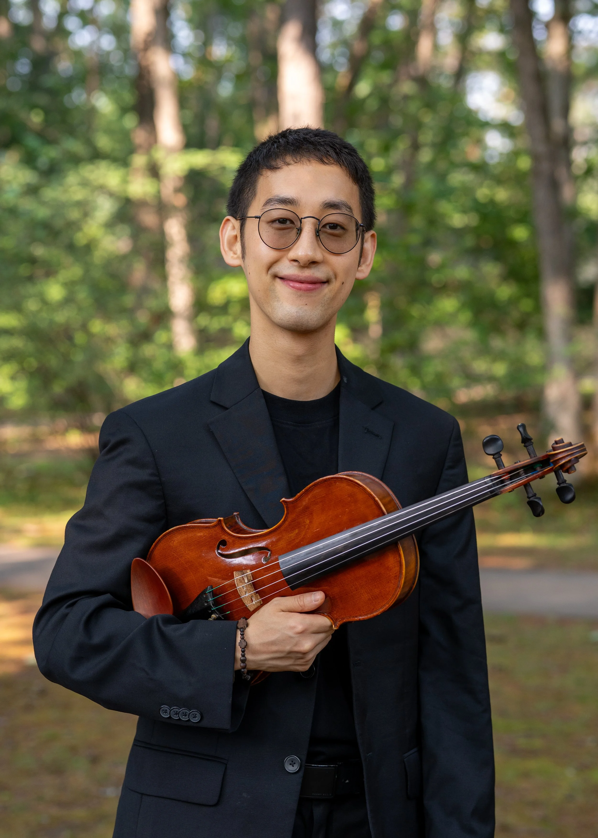 A young man with glasses and short black hair holding a violin outdoors in a wooded area, smiling at the camera.