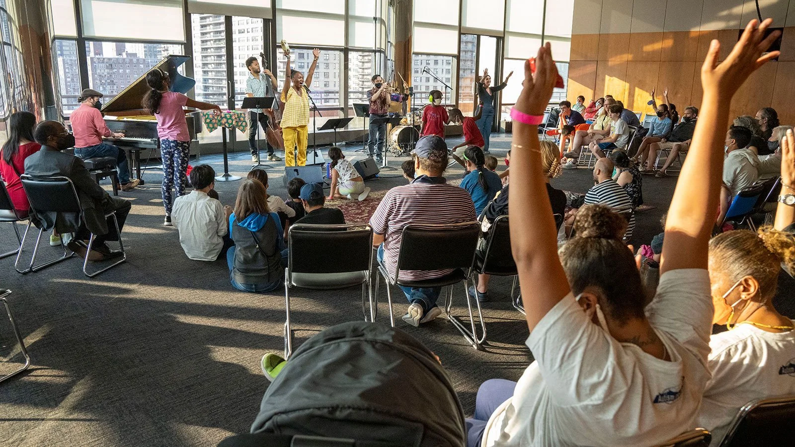 A diverse group of people attending a live musical performance in a modern indoor space with large windows and city views, some seated and some on the floor, with hands raised.