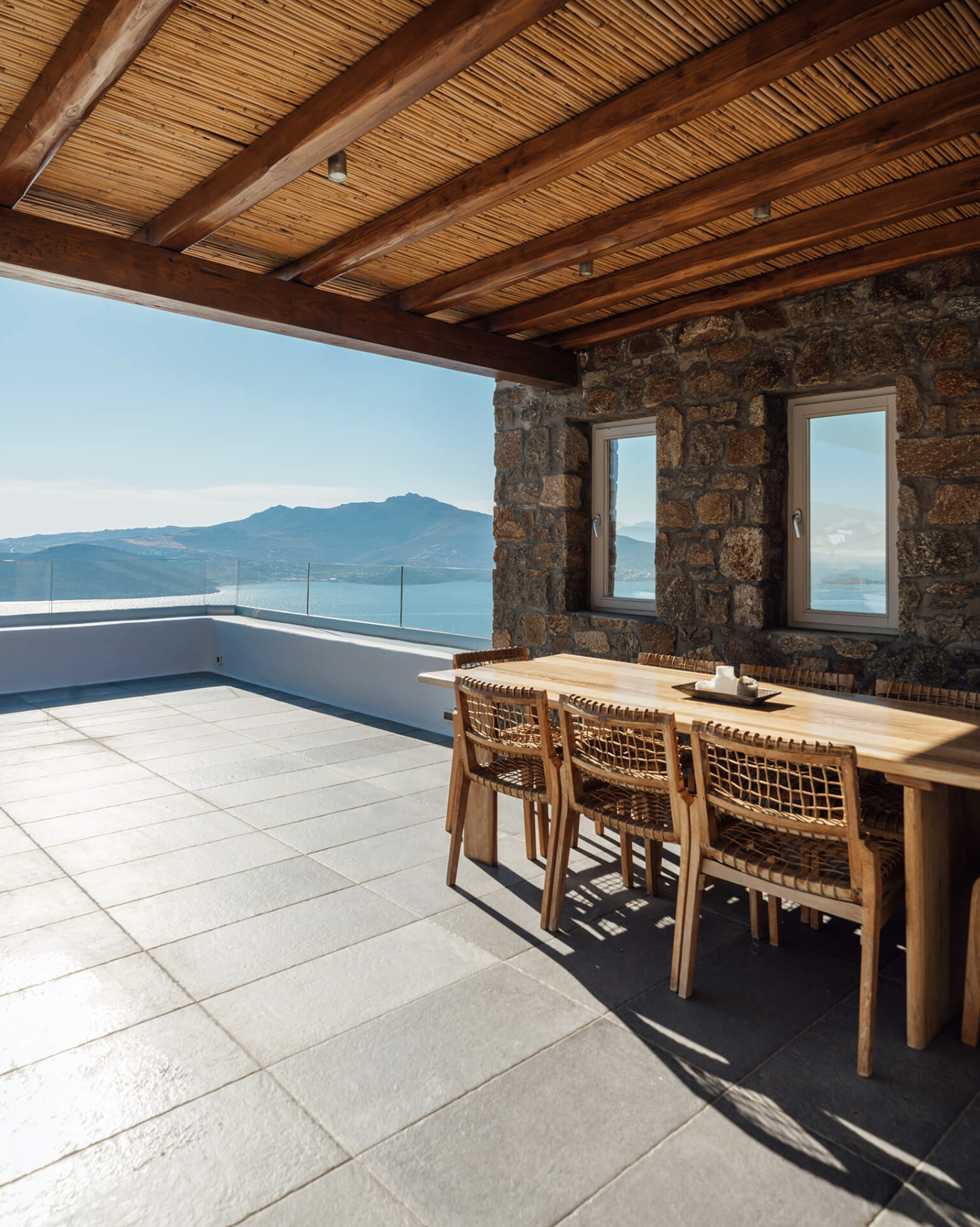 Outdoor patio with wooden dining table and chairs, stone wall with two windows, mountain and water view, cloudy sky, and tiled floor.