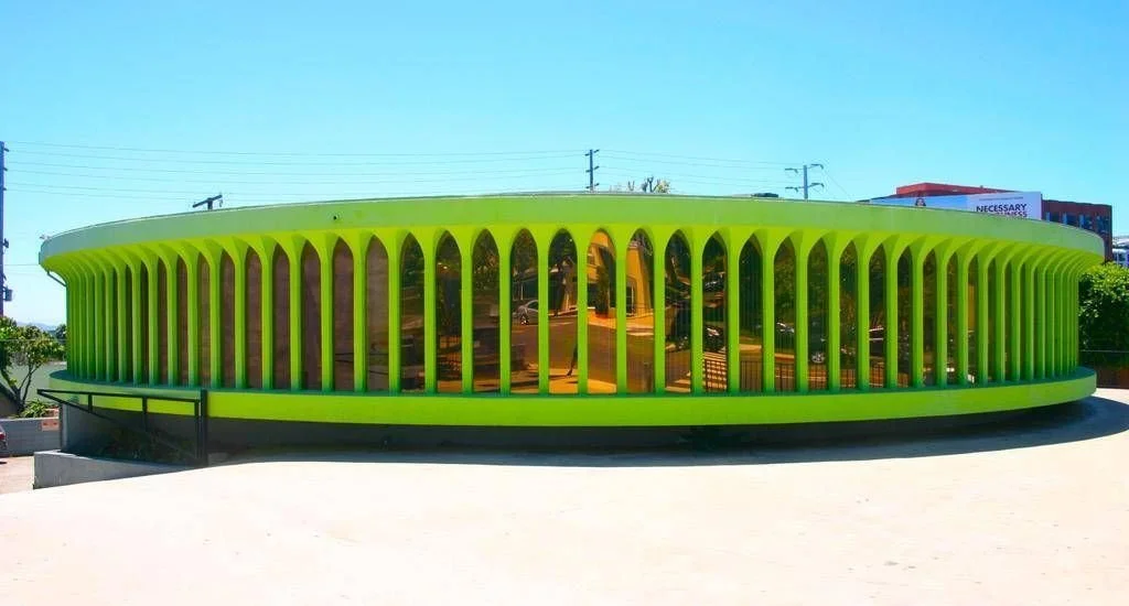 Bright yellow modern building with vertical openings and reflective windows, surrounded by pavement, under a clear blue sky.