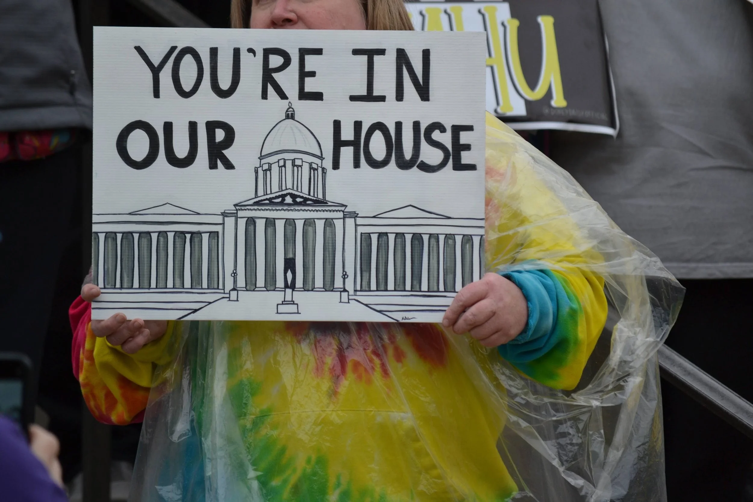 A person holding a sign that reads "You're in Our House" with a drawing of a government building, wearing a tie-dye shirt and a clear poncho.