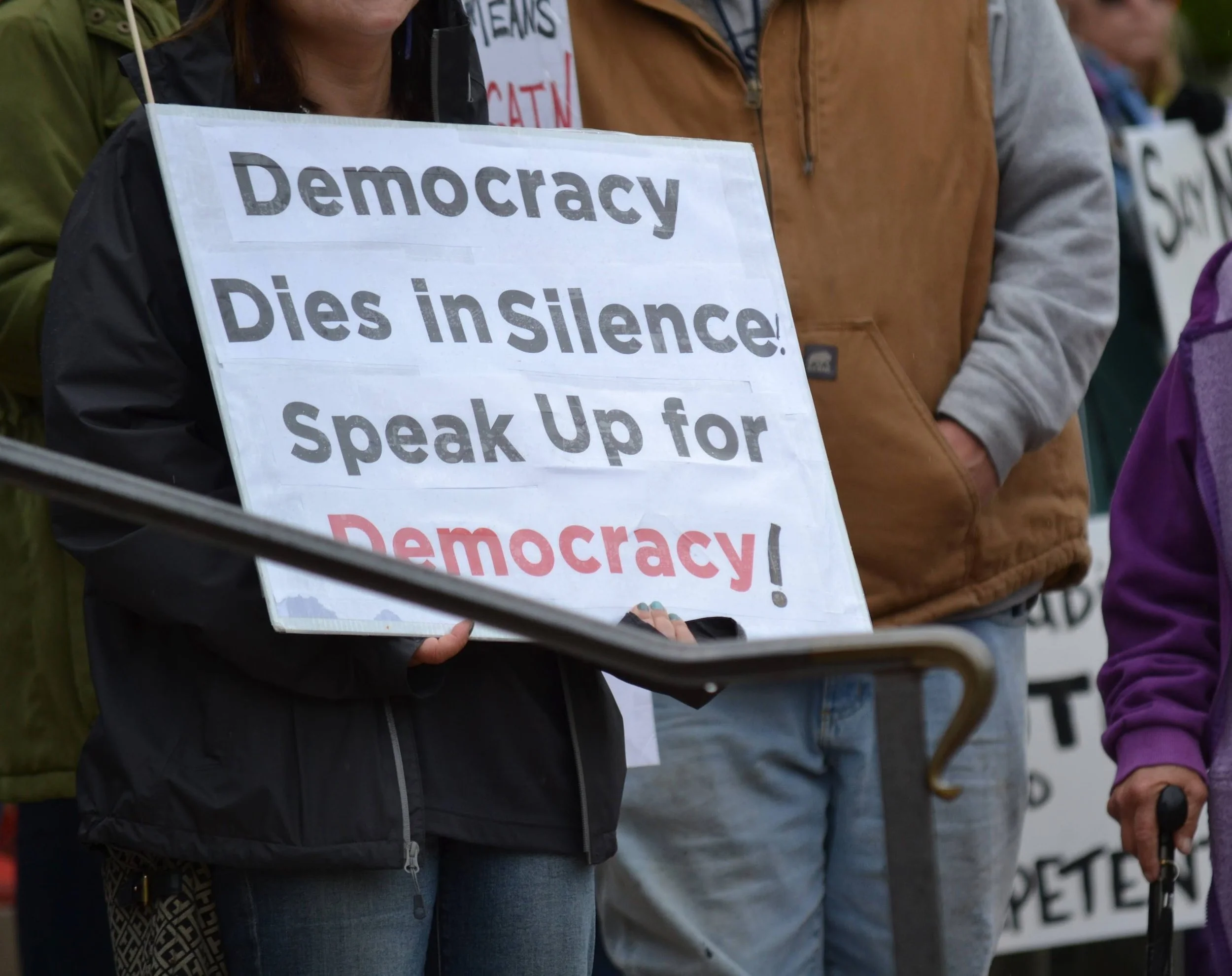 Person holding sign that reads "Democracy Dies in Silence. Speak Up for Democracy!" at a rally or protest.