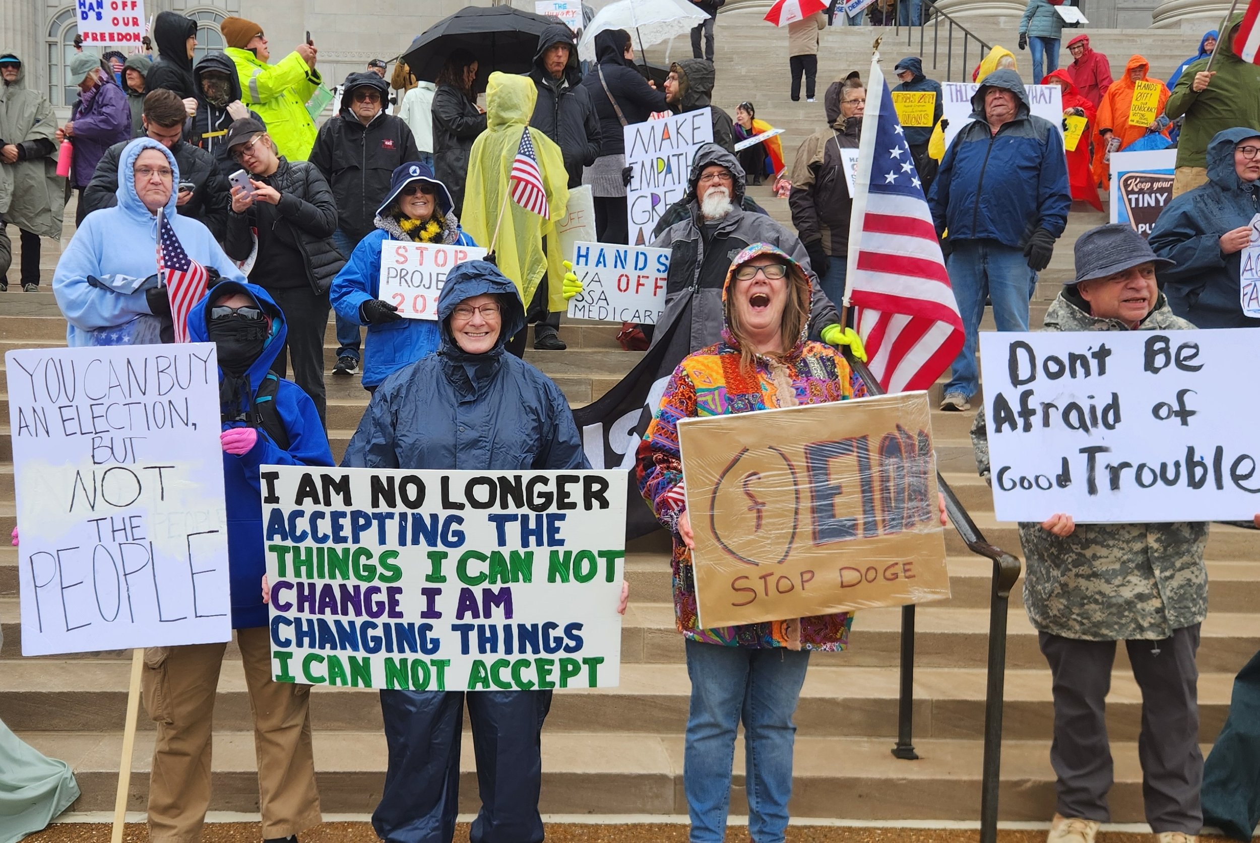 Protestors on steps holding various signs, such as "You can buy an election, but not the people" and "Don't be afraid of good trouble." Some carry American flags.