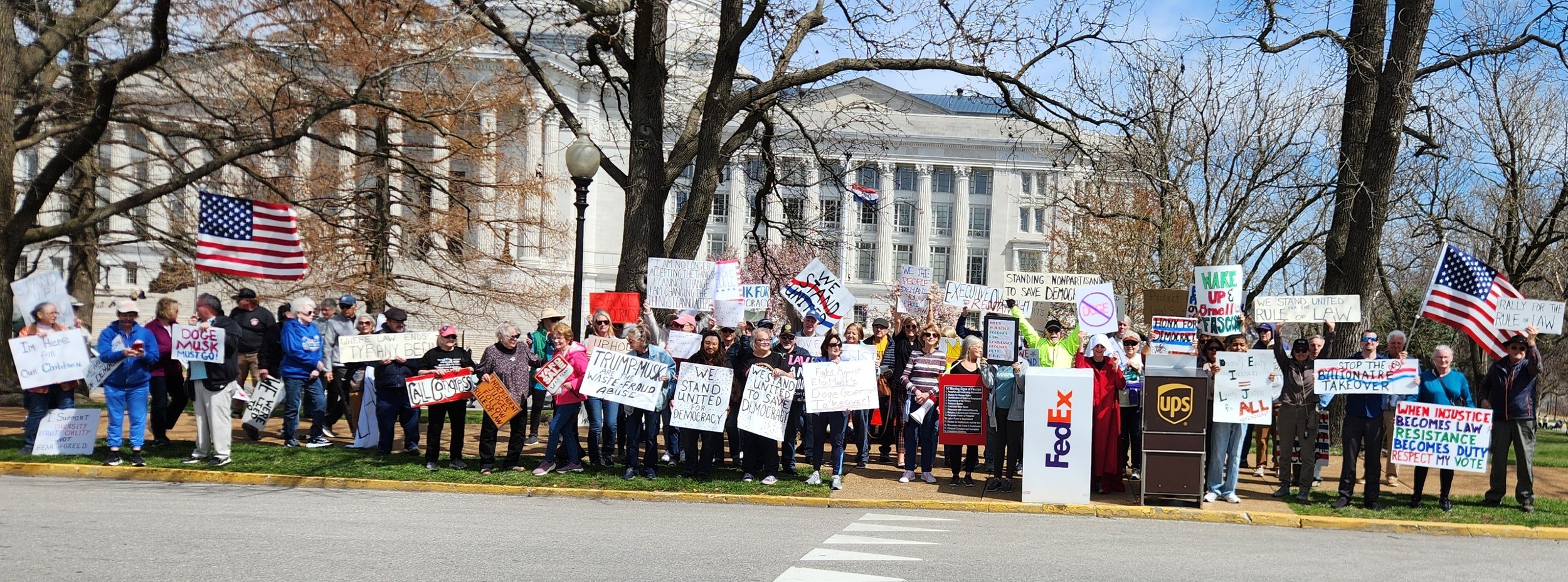 Group of people holding protest signs and American flags outside a large building.