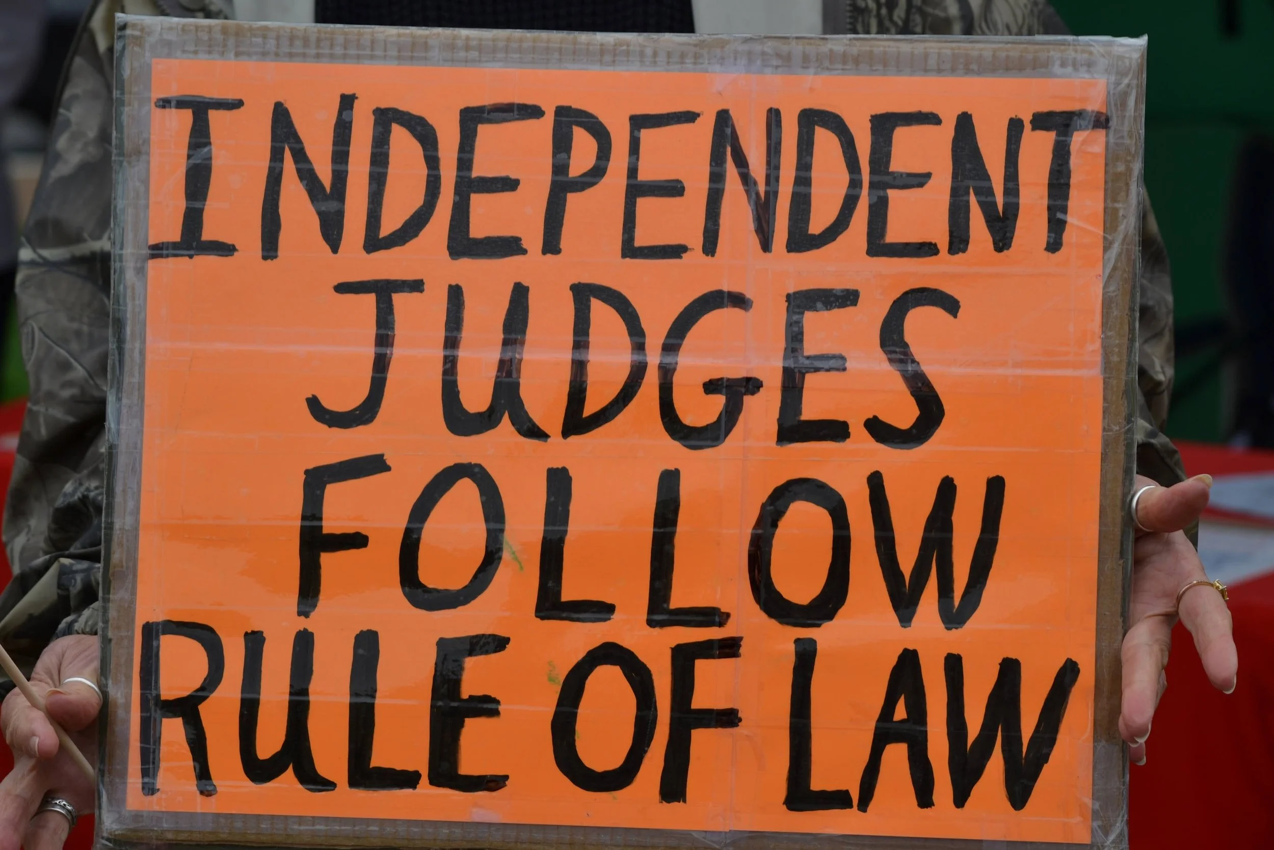 A person holding a sign with the text "Independent Judges Follow Rule of Law" in bold black letters on an orange background.