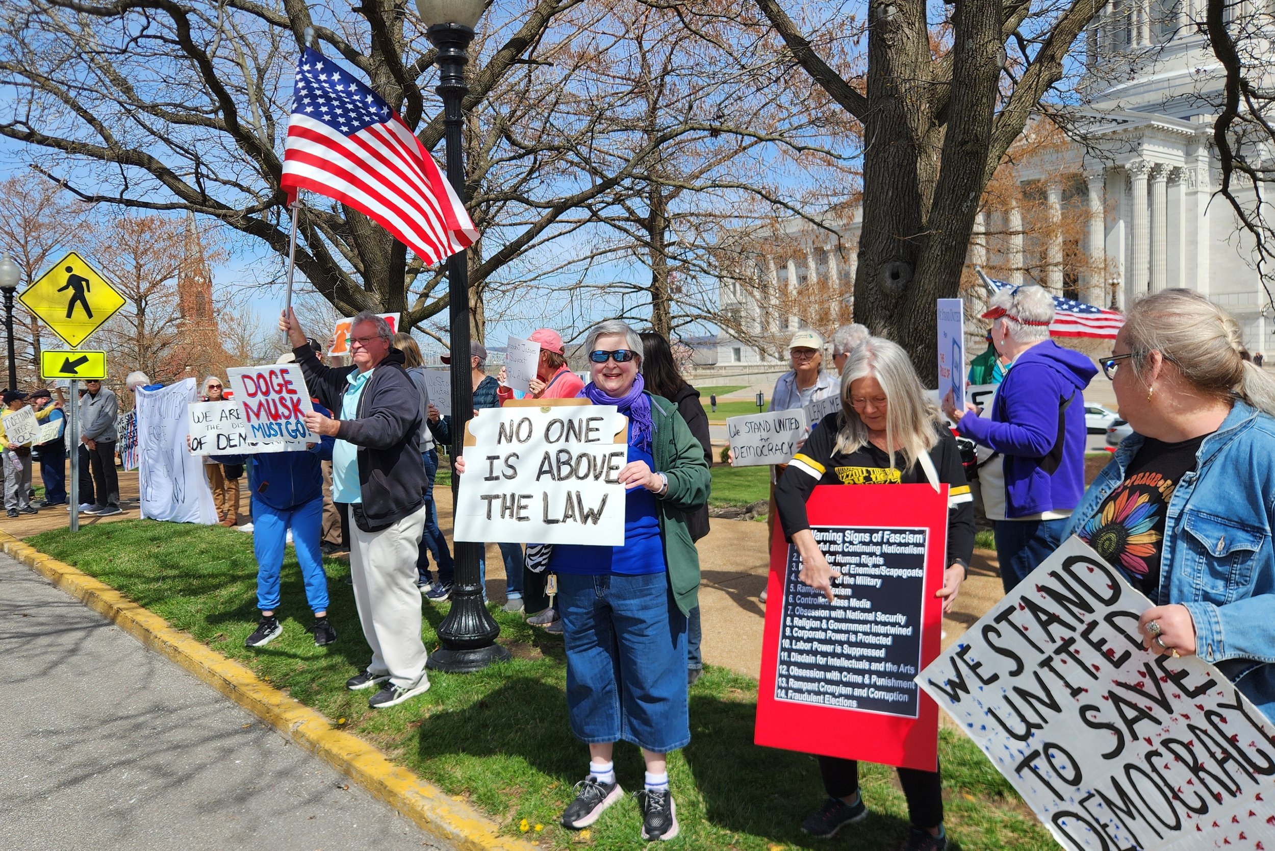 Group of protesters holding signs with messages such as "No one is above the law," standing on a street near a large building, including a person holding an American flag.