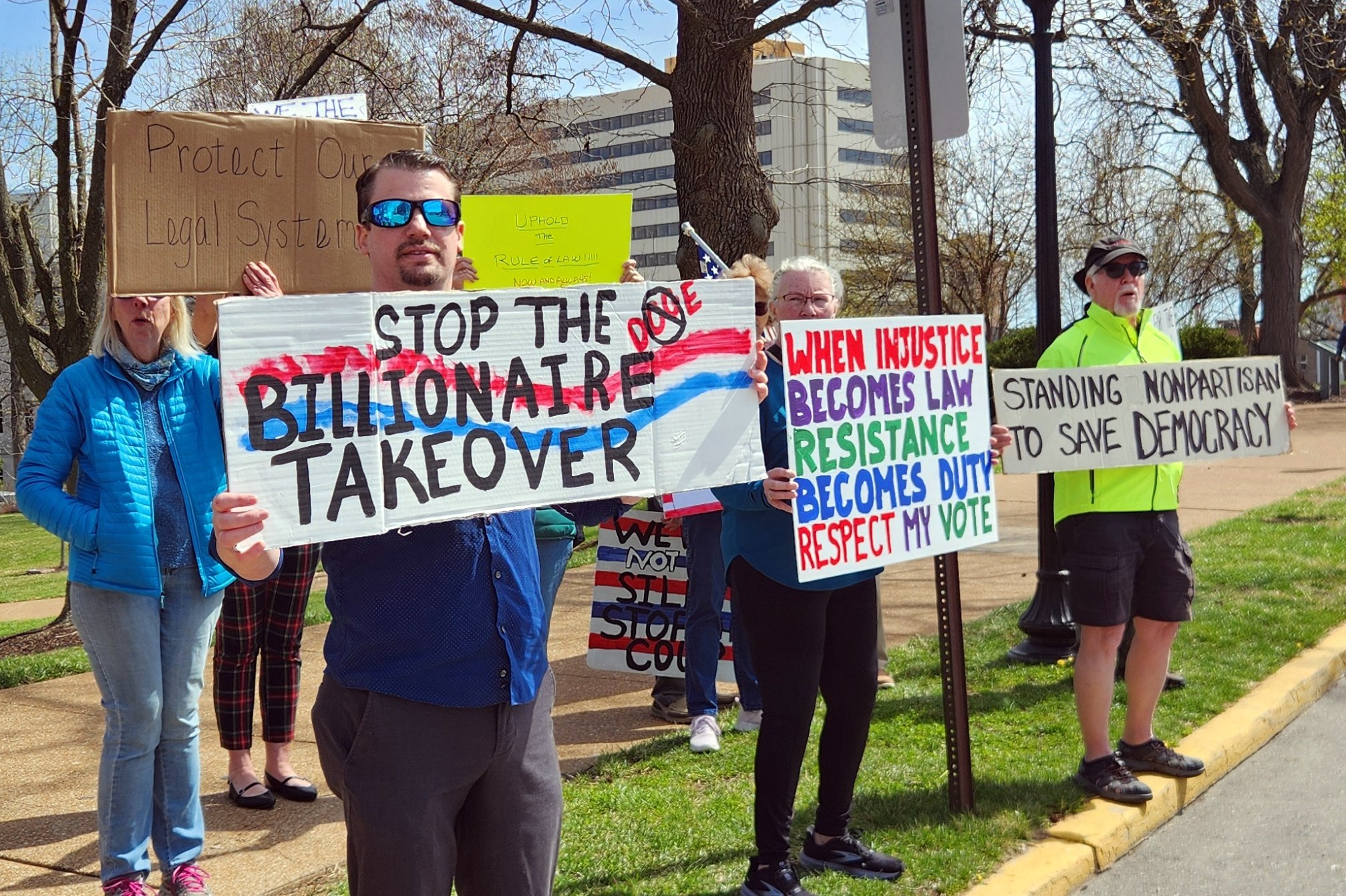 Group of protesters holding signs with messages about democracy and law, including "Stop the billionaire takeover" and "Standing nonpartisan to save democracy," on a sidewalk with buildings and trees in the background.