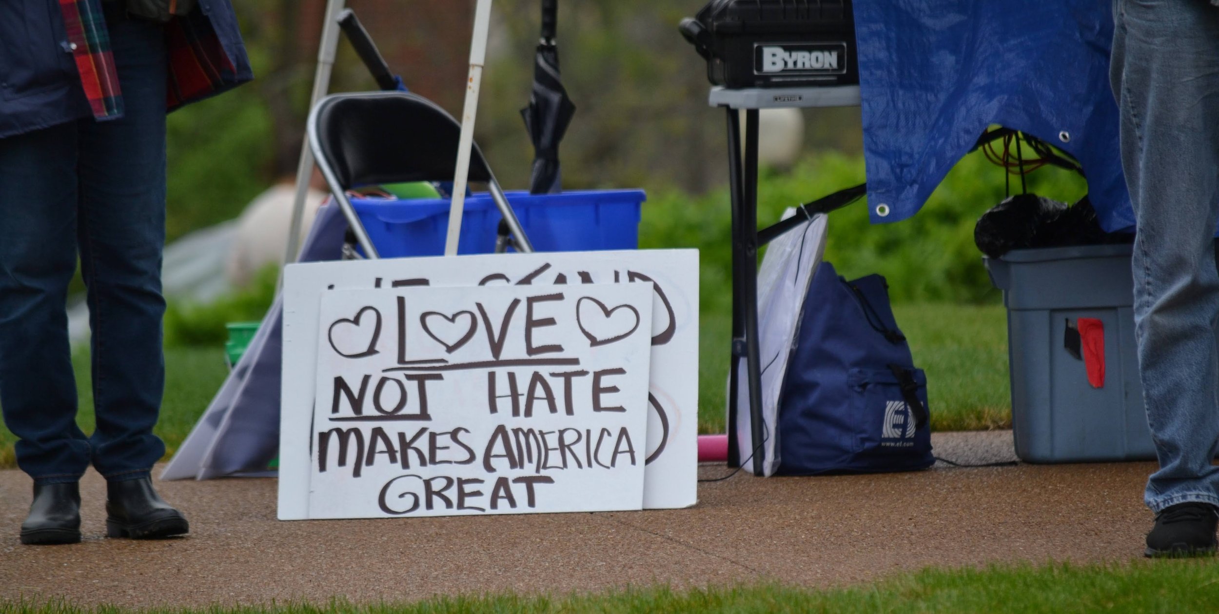 Protest sign on the ground reading 'Love not hate makes America great' partially obscured by other items, blue storage bins, and people standing nearby.