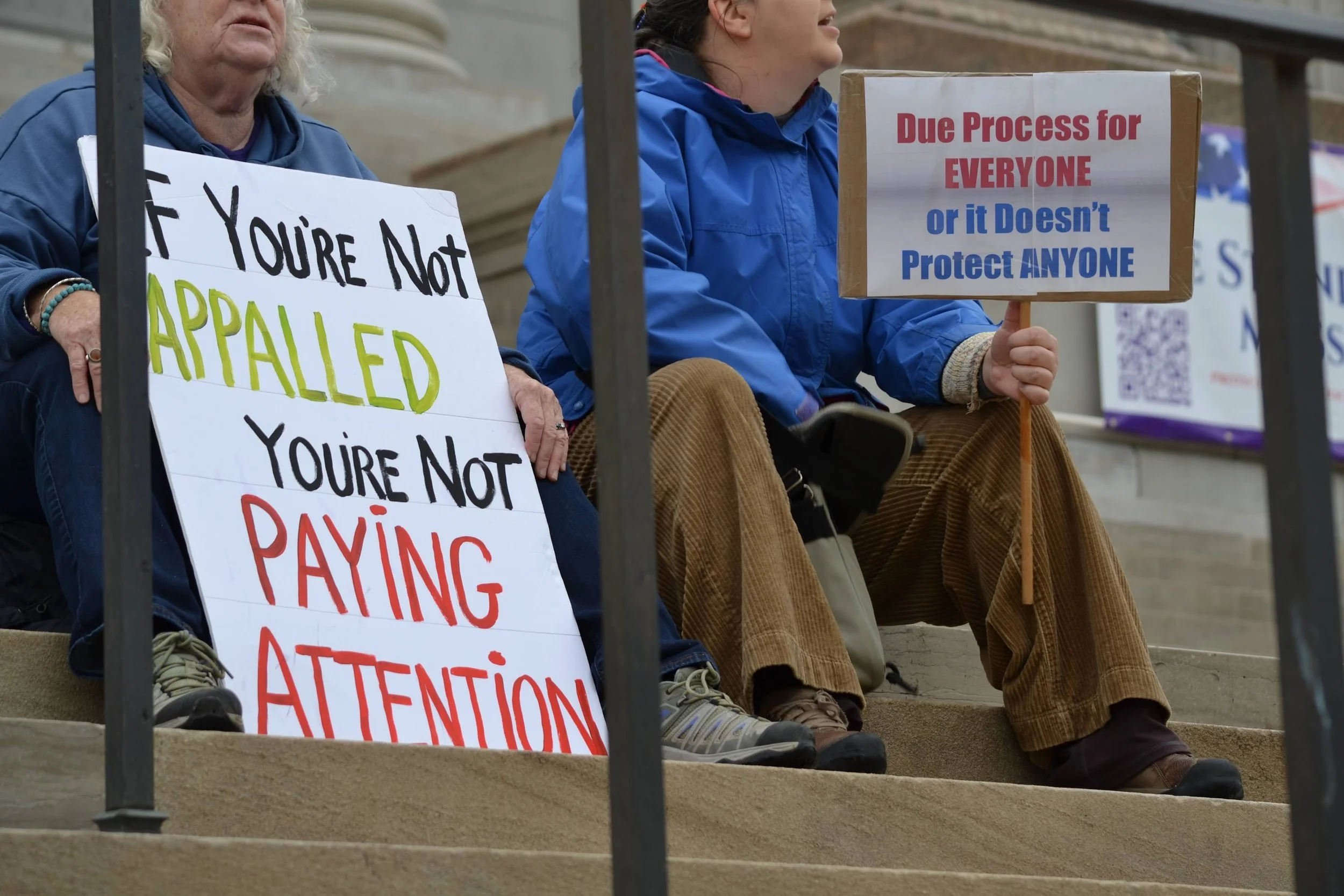 Protesters sitting on steps holding signs, one reads "If you're not appalled, you're not paying attention," and "Due process for everyone or it doesn't protect anyone."