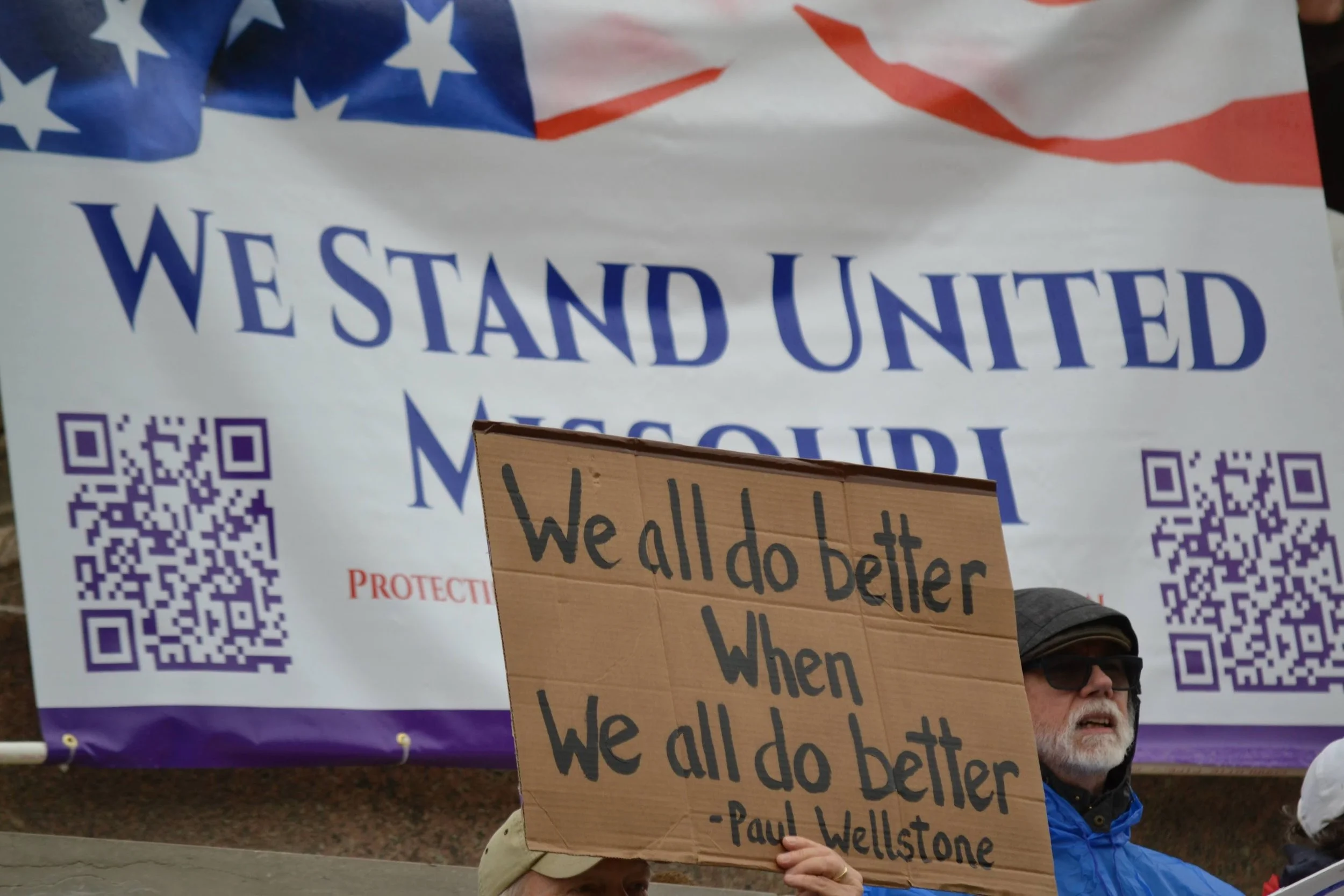 Protest sign reading, "We all do better when we all do better" held at a rally in front of a banner with American flag elements and the text, "We Stand United Missouri."
