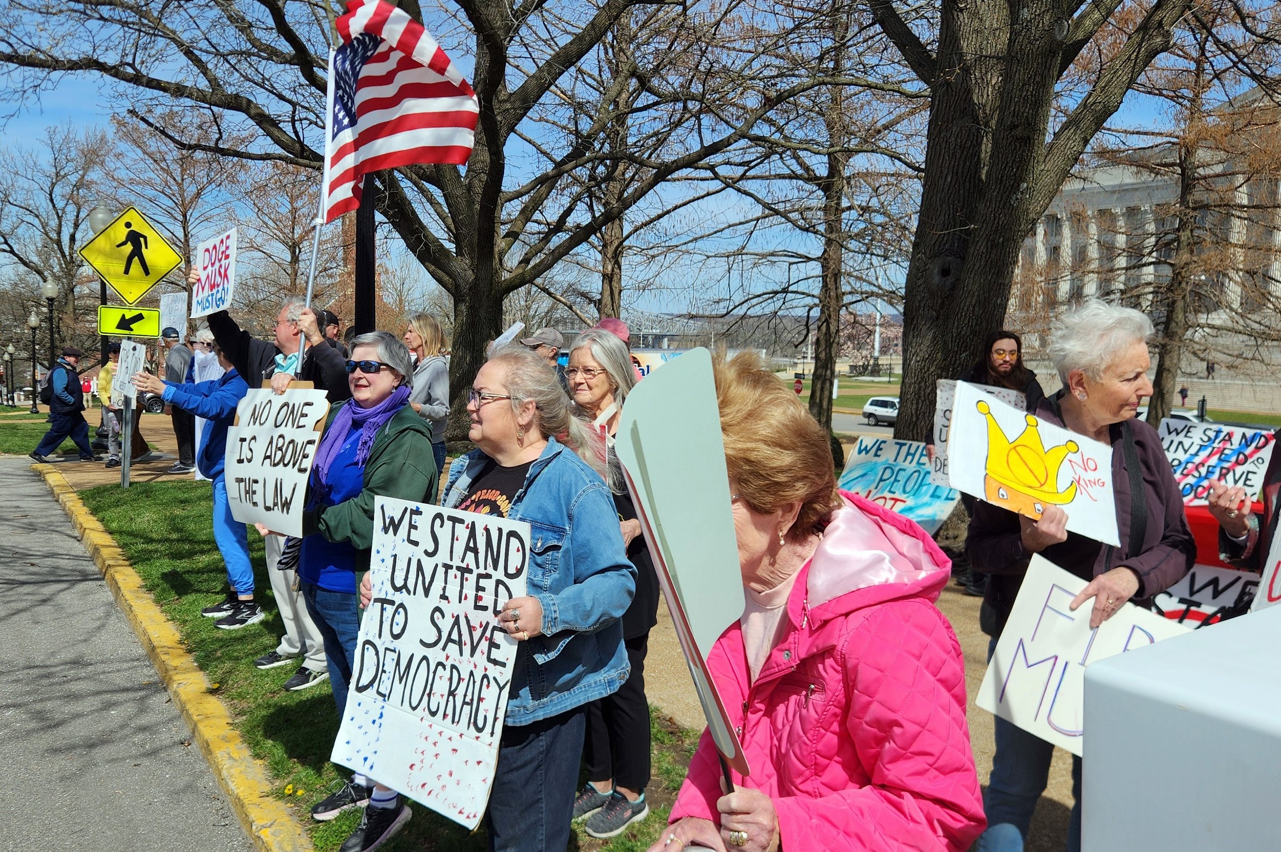 A group of people holding protest signs, including phrases like "No one is above the law" and "We stand united to save democracy," with an American flag and a crosswalk sign on a tree-lined street.
