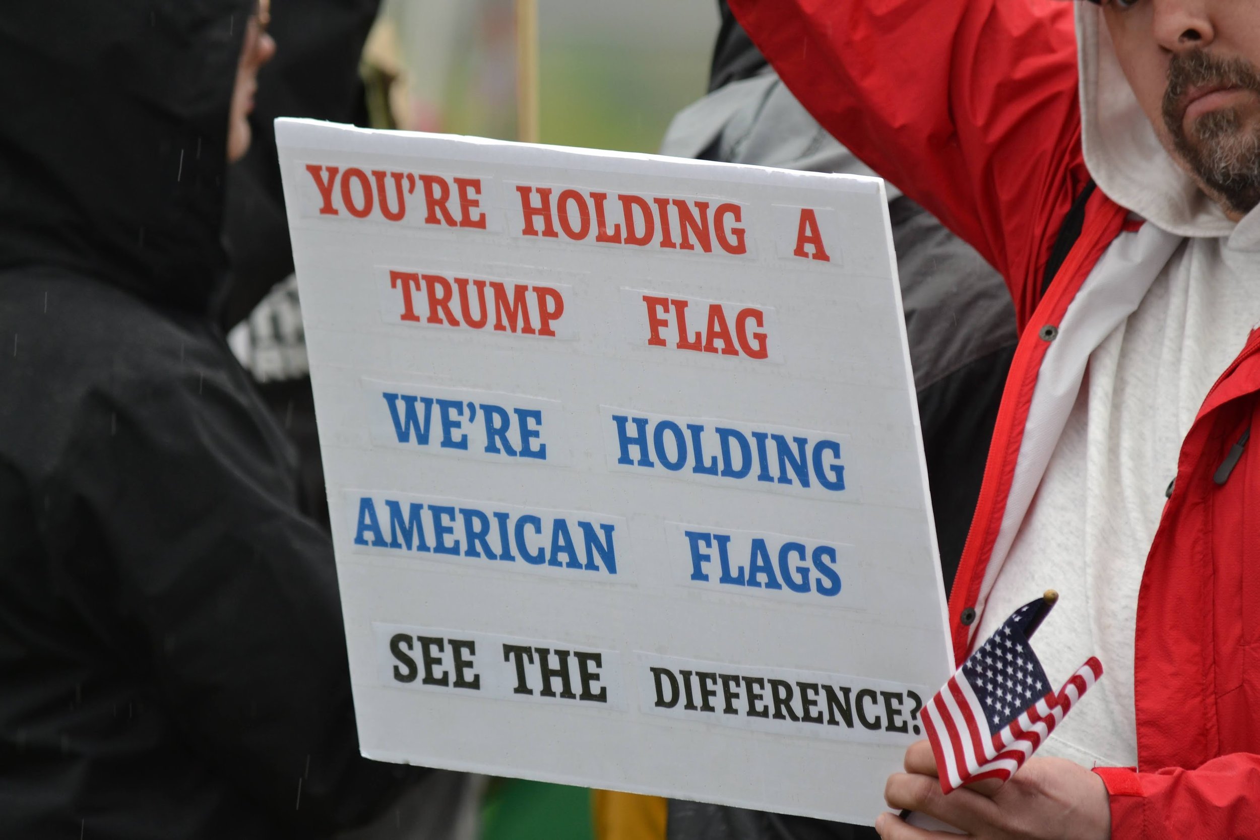 Person holding sign with text contrasting 'Trump flag' and 'American flag', holding a small American flag.