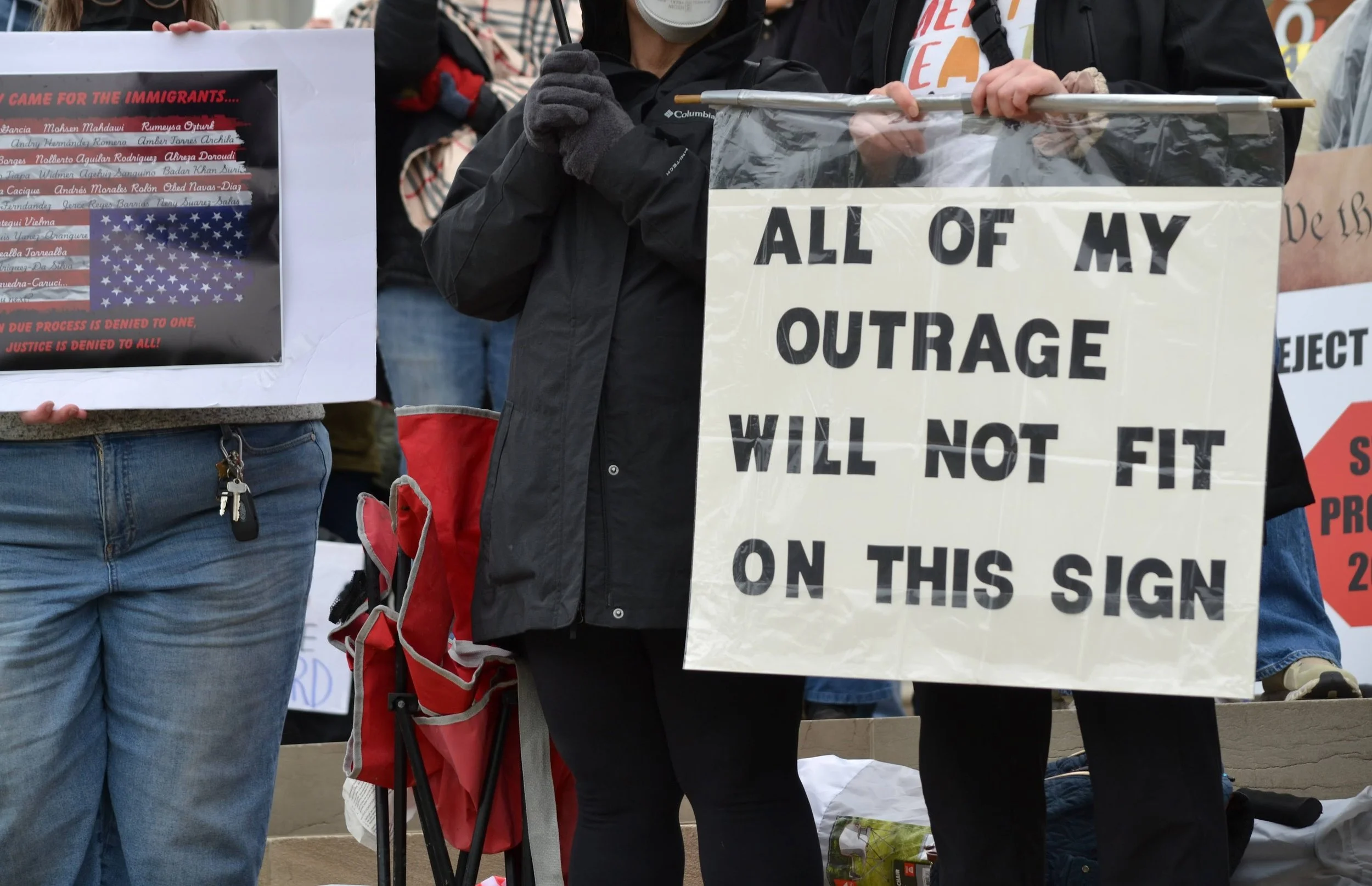 People holding protest signs about immigration, one stating "All of my outrage will not fit on this sign."