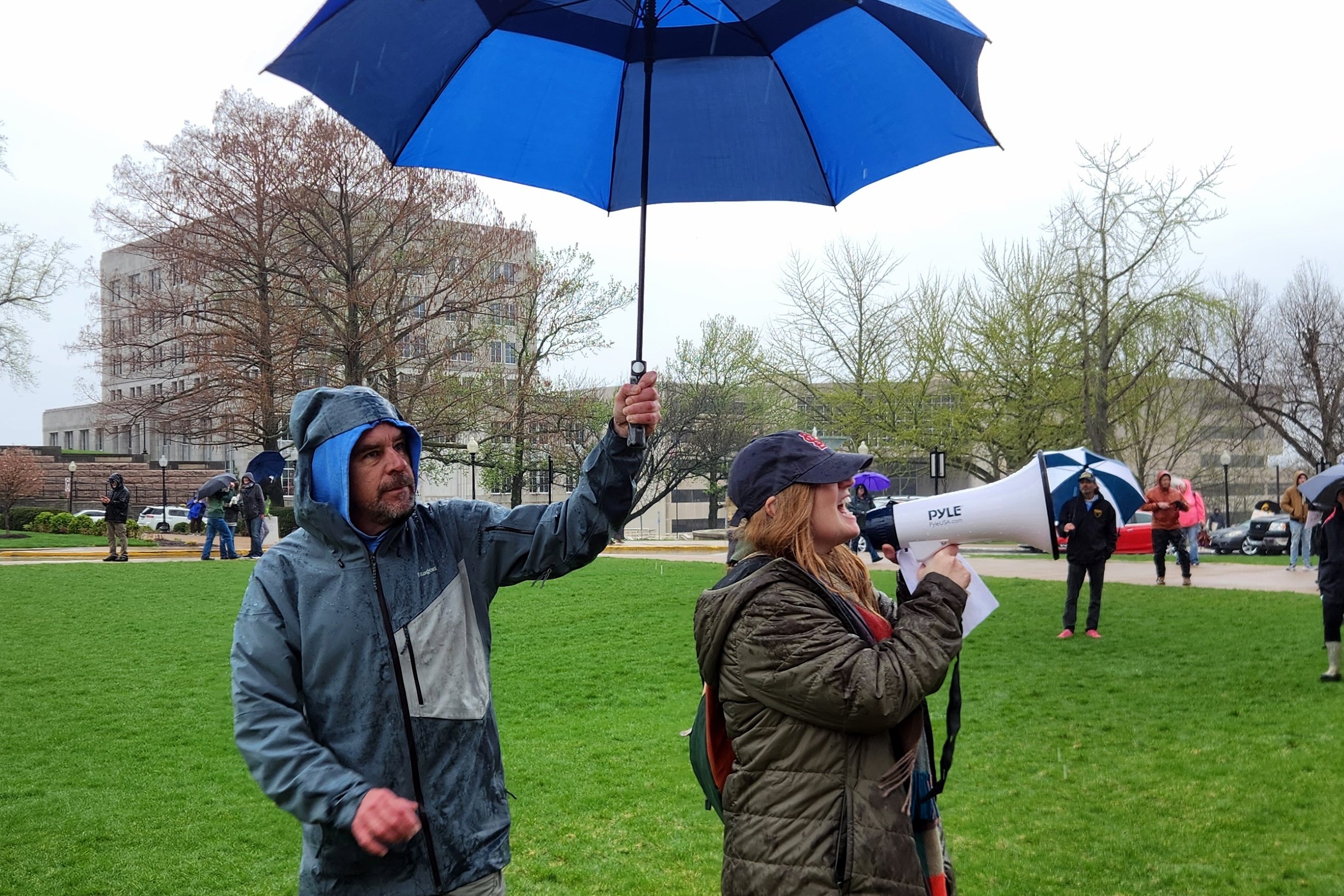 Two people in a park on a rainy day. One person holds an umbrella, while the other speaks into a megaphone. A public building and several other people with umbrellas are visible in the background.