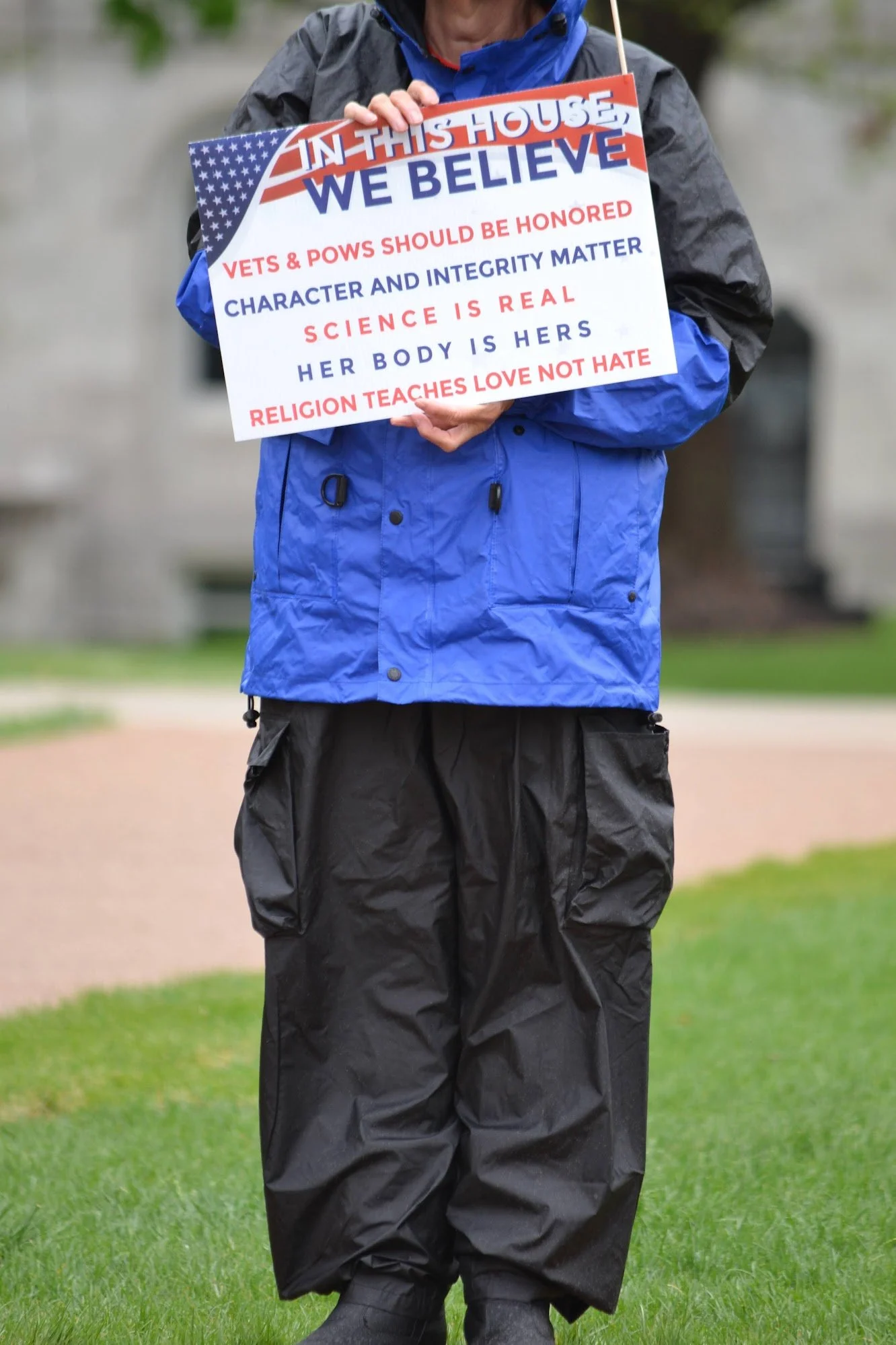 Person holding a sign with messages about beliefs, wearing a blue jacket and black pants, standing on grass.
