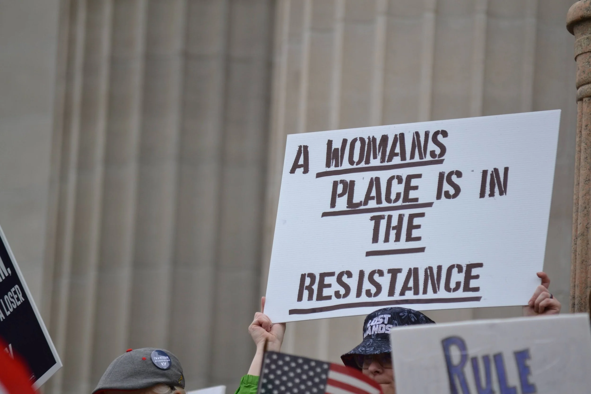 Protester holding a sign that reads, "A Woman's Place Is in the Resistance," in front of a building with columns.