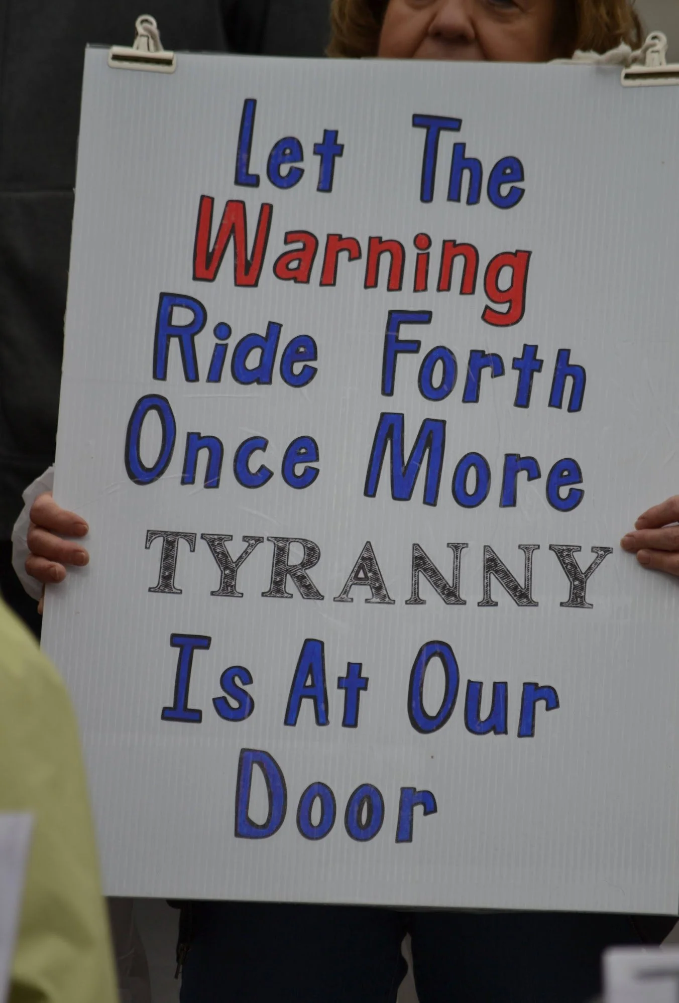 A protest sign reading 'Let The Warning Ride Forth Once More TYRANNY Is At Our Door' held by a person.