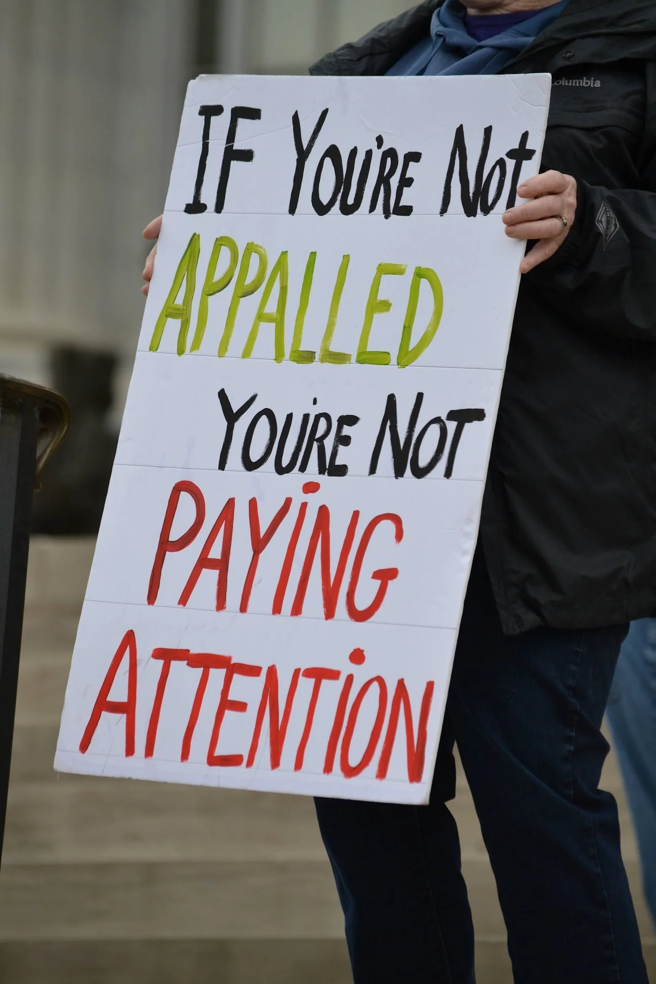 Person holding a protest sign reading, 'If You're Not Appalled You're Not Paying Attention.'