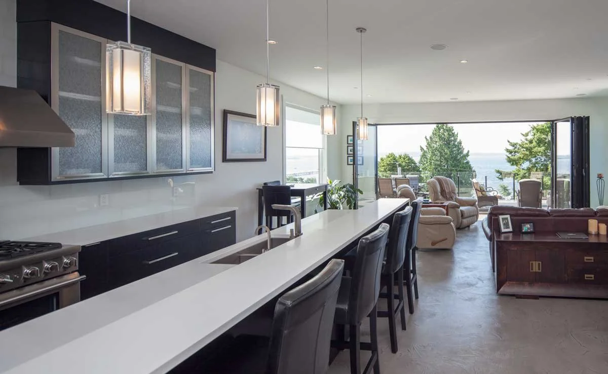 Modern open-plan kitchen and living room with large windows and sea view, featuring a long kitchen island, bar stools, pendant lights, and outdoor seating area. Converge Construction