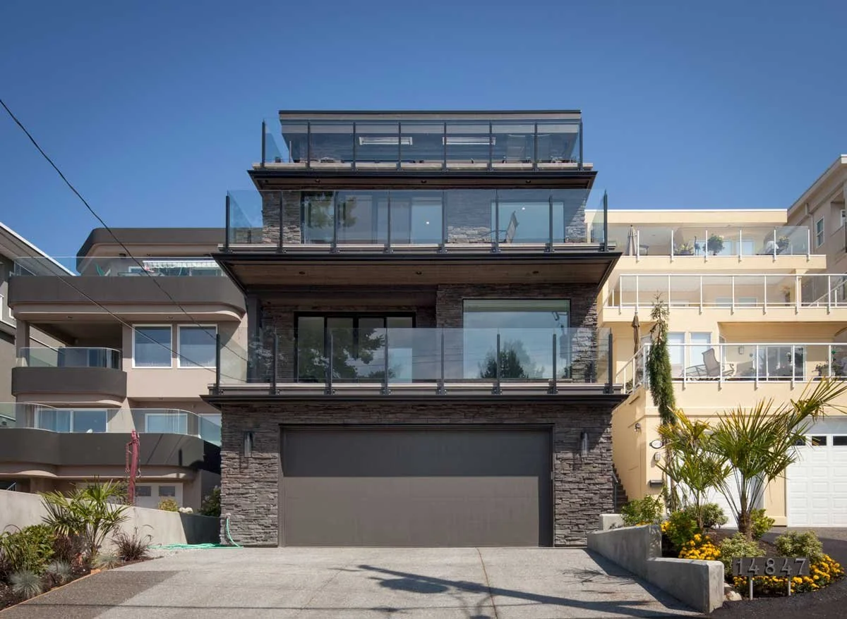 Modern three-story residential house with glass balconies, stone facade, and a large garage, surrounded by landscaped plants, under a clear blue sky.