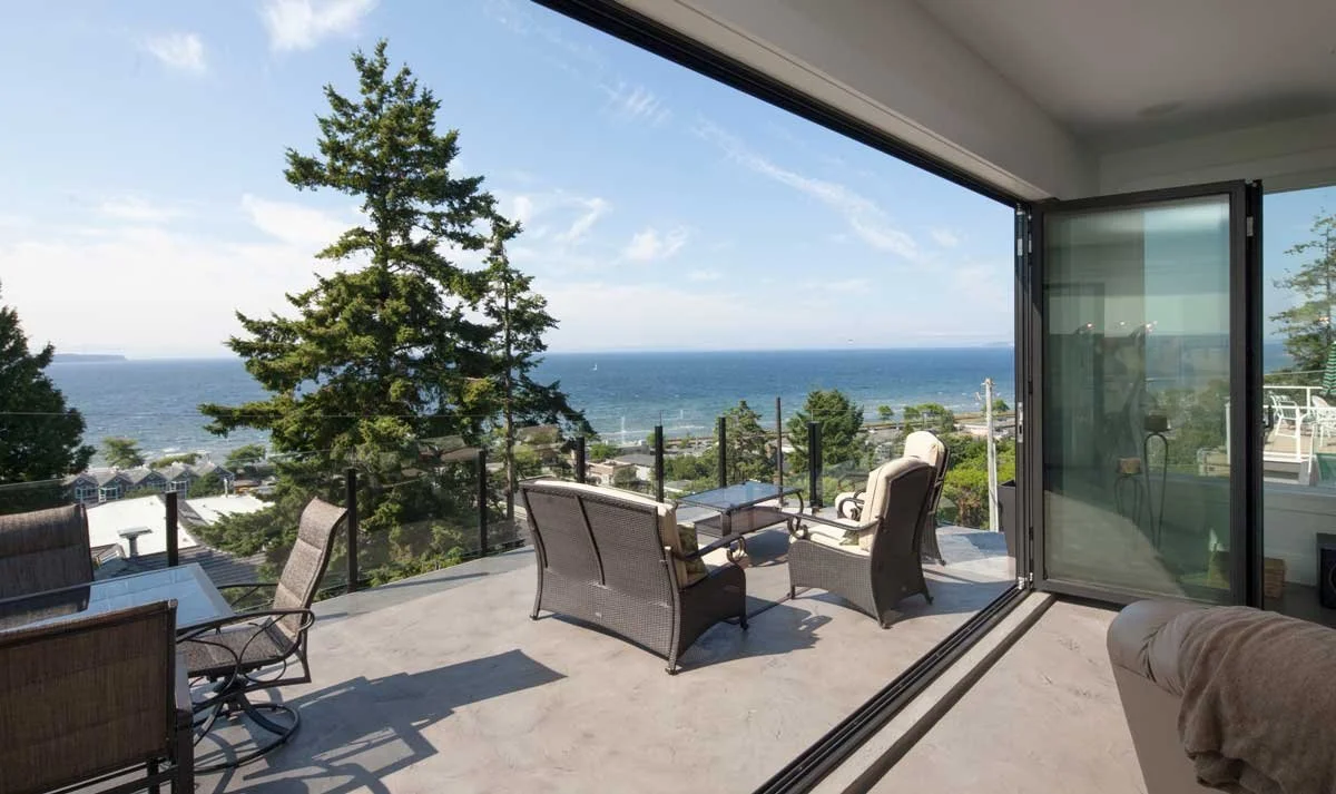 Luxury patio with glass railing overlooking ocean and trees; outdoor seating area with chairs and table.