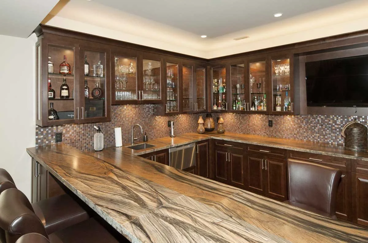 Home bar with marble countertop, wooden cabinets, glass shelving displaying bottles, a small sink, stools, and a TV.