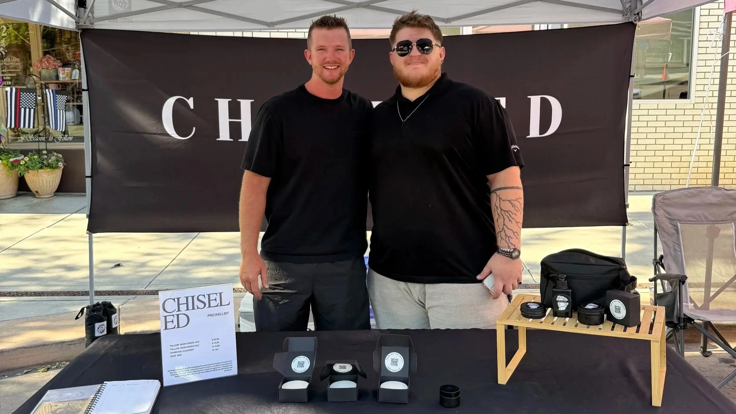 Two men standing behind a table at an outdoor stall, smiling at the camera. One man wears sunglasses and a black t-shirt, the other wears a black shirt and has tattoos on his arm. The table displays product packaging and a pricing list, with a large black banner in the background.