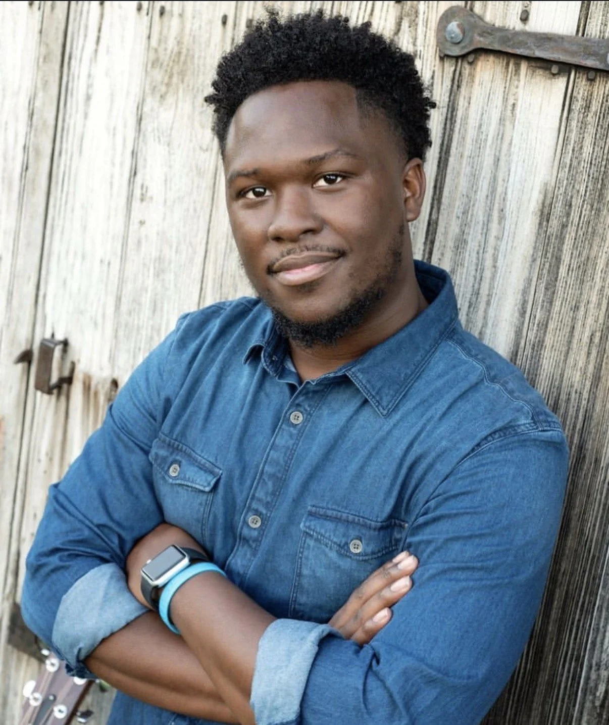 A young man with short curly hair and a beard, wearing a gray zip-up sweater, posing against a blue background.