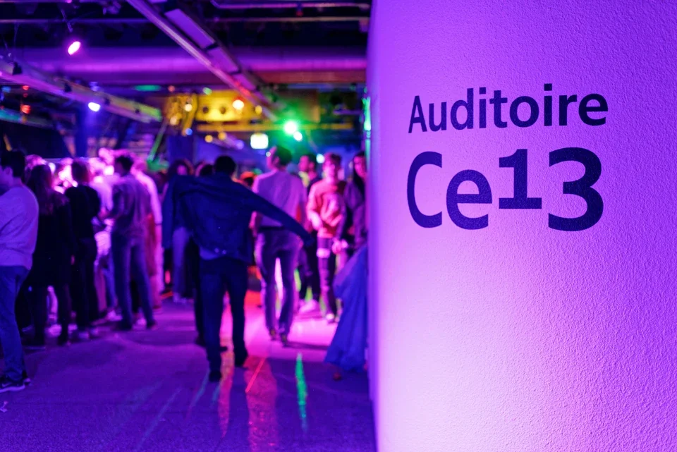 Une foule de personnes à l'intérieur d'un espace avec éclairage coloré, près d'un mur avec une inscription 'Auditorie Ce13'.