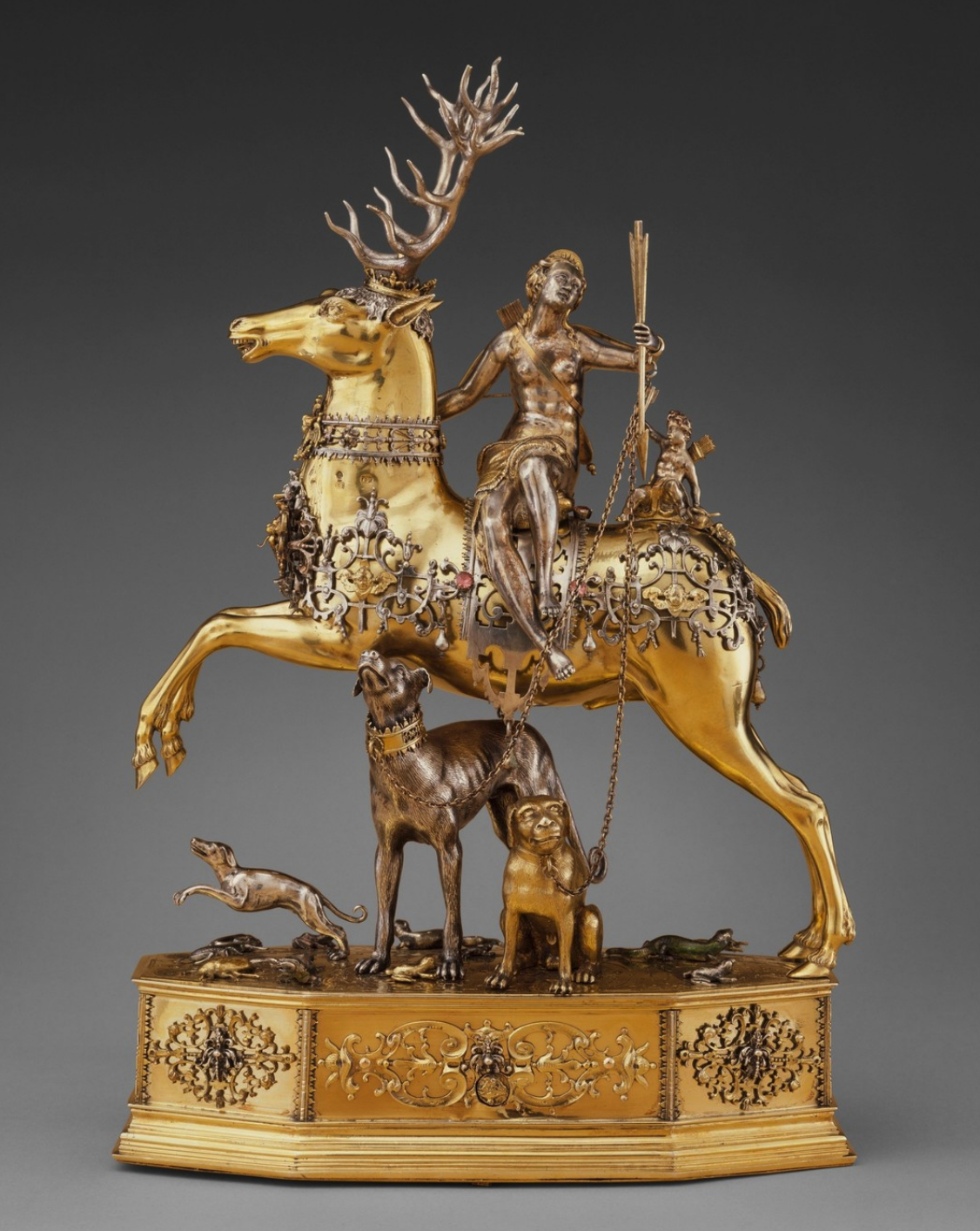 A partially nude female figure sits sideways, glancing upwards with an arrow in hand atop a lavishly decorated golden stag, while a small cupid figure sits behind her.