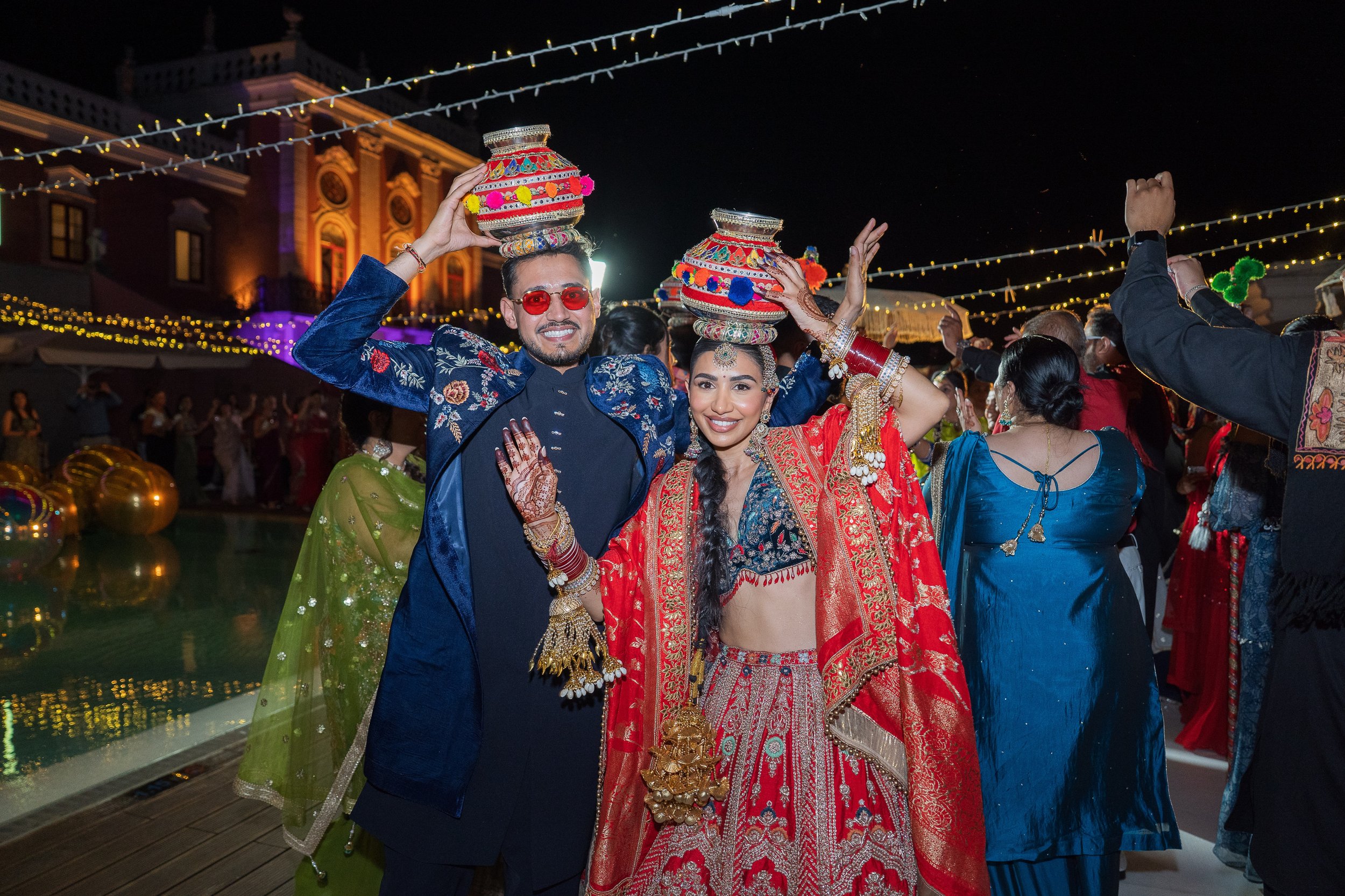 Bride and groom wearing festive attire carrying decorated pots on their heads during an Indian wedding celebration at an outdoor Algarve venue at night.