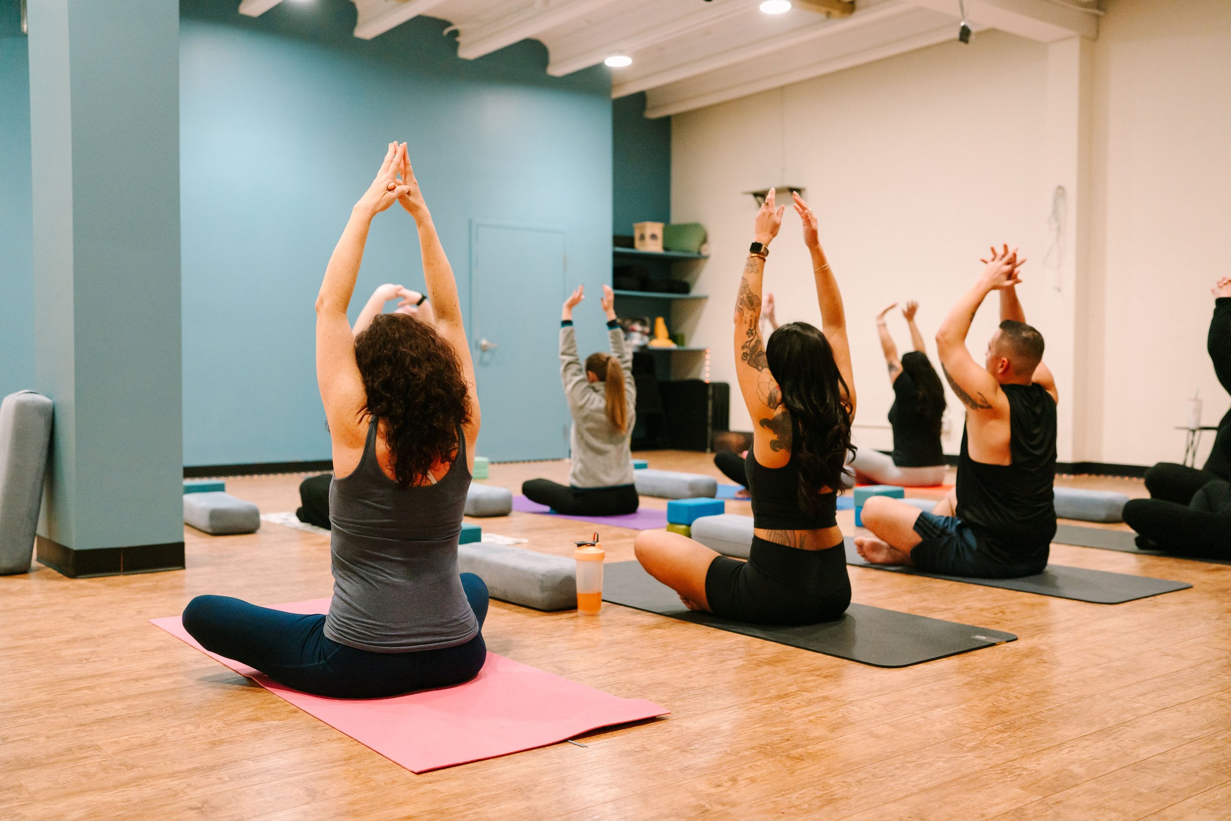 People practicing yoga in a studio, sitting on yoga mats with arms raised above their heads in a seated position.