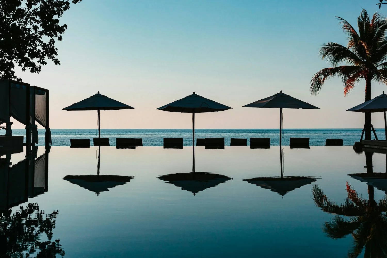 Sunset view of a beachfront pool area with five umbrellas and chairs facing the ocean, reflected in the pool, with palm trees on the right and silhouettes of trees on the left.