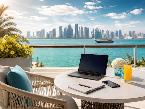 A balcony with a white table and cushioned chair overlooking the ocean and a city skyline in the distance. On the table are a laptop, a notebook, a pen, a smartphone, a glass of orange juice, and a bouquet of white flowers.