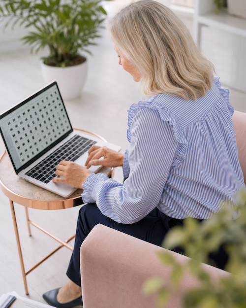 An older woman with gray hair working on a laptop at a small round table in a bright living room, with a large potted plant behind her.