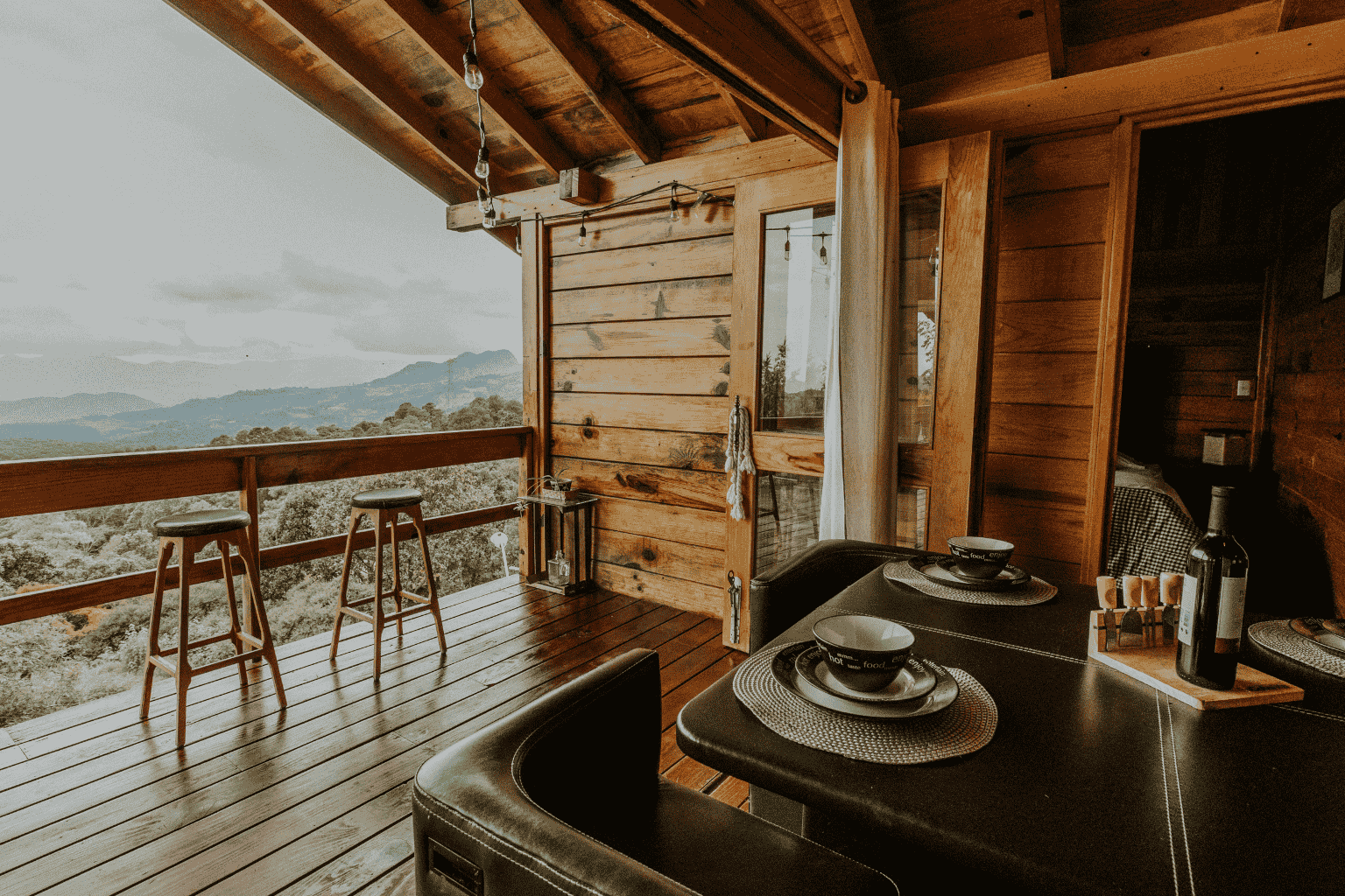 Interior view of a wooden cabin with an open balcony overlooking a mountain landscape, with two wooden stools, a lantern, and a dining table set with black bowls and plates.
