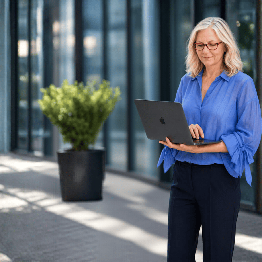 An middle aged woman with blonde hair and glasses looking at a laptop outdoors in front of a modern glass building and potted plant.