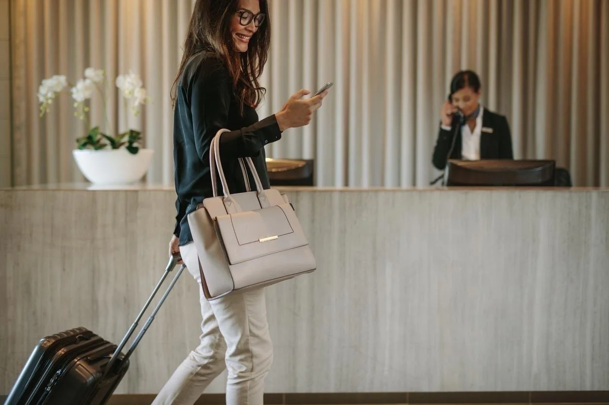 A woman with glasses and a beige handbag checking her phone at a hotel reception desk, pulling a rolling suitcase, with a staff member on the phone in the background.